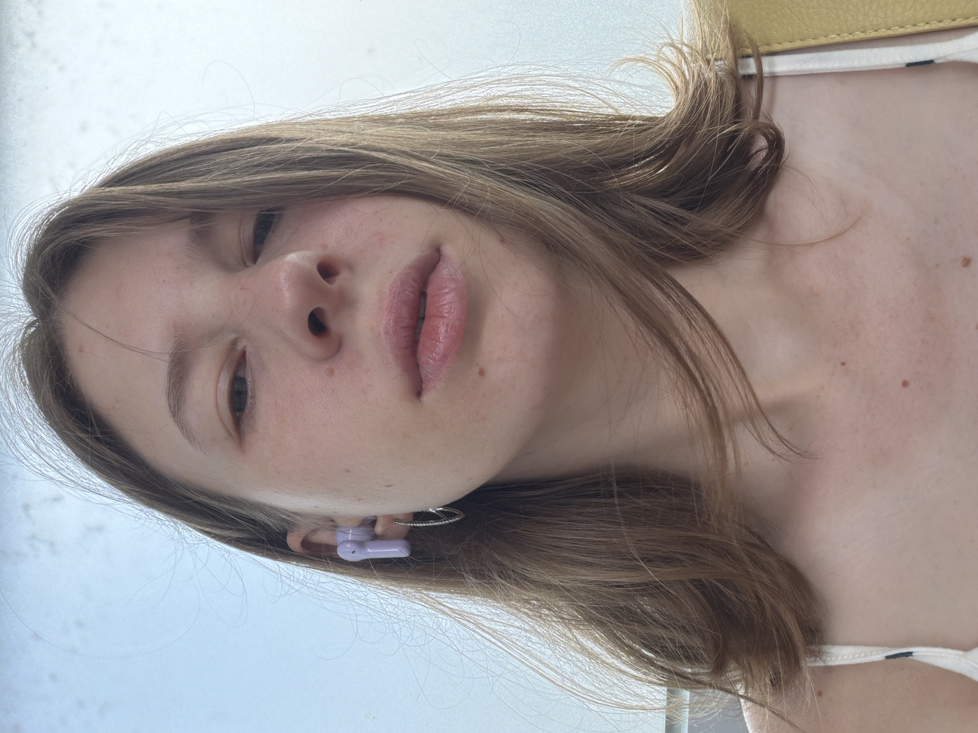 A young woman is captured in a close-up, eye-level, selfie-style photograph taken outdoors against a bright, overcast sky. She has light brown hair, parted in the center, with strands falling around her face. She is wearing a white tank top with a subtle pattern. In her left ear, she has a light purple earbud and a small silver hoop earring. Her skin has a light complexion with some freckles. Her lips are slightly parted, and her gaze is directed upwards towards the camera. The background is a blurry expanse of sky, suggesting an open, outdoor environment. No other individuals or specific landmarks are visible in the frame.