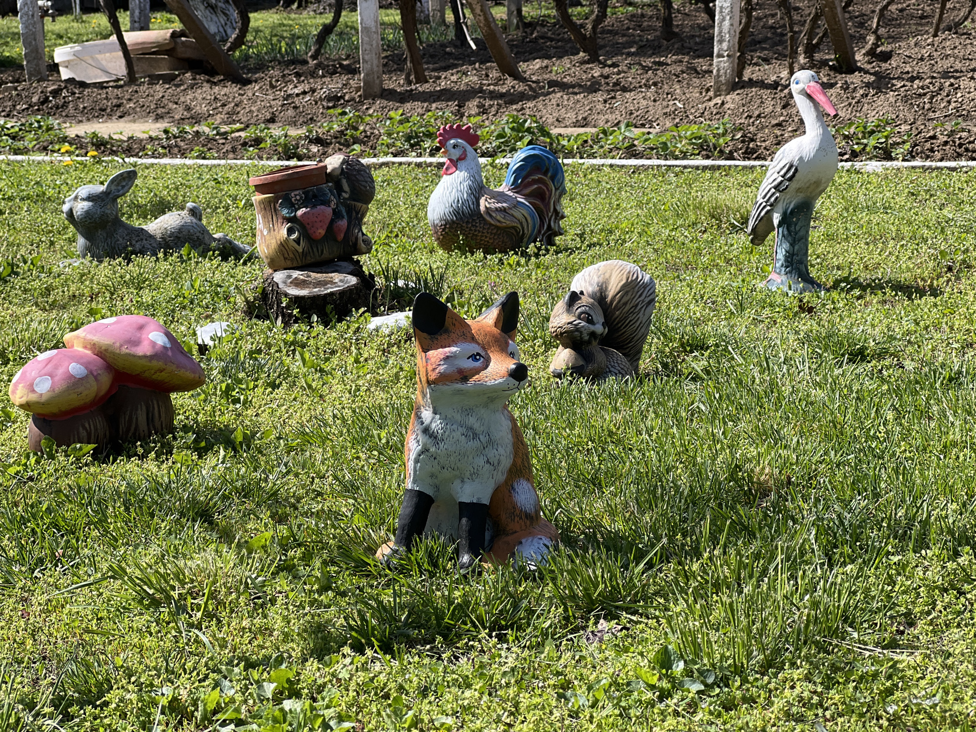 The image depicts a garden scene in what appears to be Lyubenovo, Bulgaria, based on geographic assumptions. Several ceramic garden ornaments are scattered across a lawn of green grass mixed with clover and weeds. The ornaments include a lying rabbit, a container on a stump, a colorful rooster, a fox, a squirrel, a bird, and a cluster of mushrooms. In the background, there is a tilled patch of earth with young plants, delineated by a white line. Tree trunks and branches are also visible in the background. The lighting suggests a sunny day. No people or actions are visible in the scene.