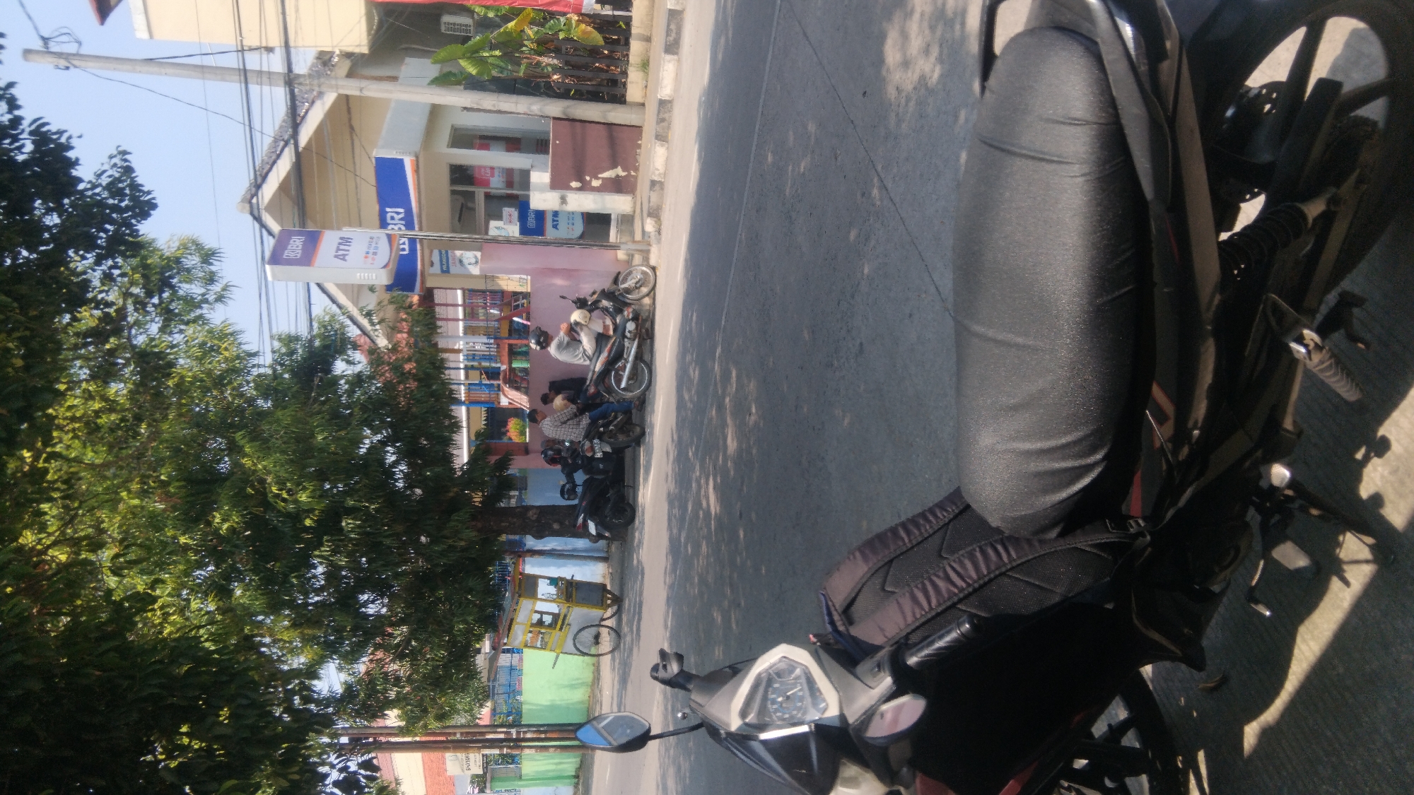The image captures a street scene in Proyonangg, Indonesia, on a sunny day. In the foreground, the seat and part of a black motorcycle are visible. In the middle ground, several other motorcycles are parked along the side of the road. Two individuals are seated on one of the motorcycles, facing away from the camera. Another person is standing next to them, and two more individuals are seated on another motorcycle further down. Behind the motorcycles, a food cart with a yellow awning is visible. To the right, a building with a blue BRI bank sign and an ATM is situated. A large tree with lush green foliage shades a portion of the scene. The road surface appears to be asphalt, with a sidewalk and curb separating it from the building. The background features more buildings and a clear blue sky.
