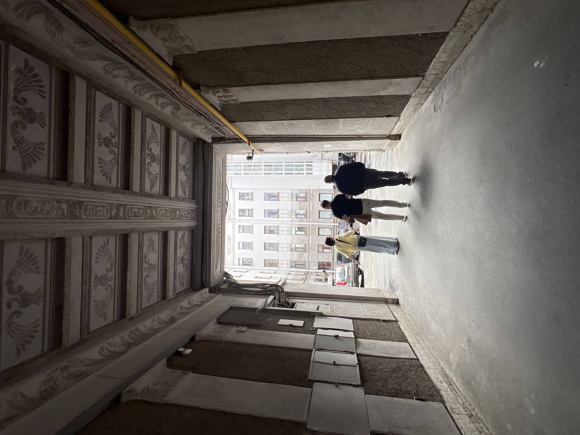 The image shows an overcast day in Saint Petersburg, Russia, likely in the afternoon. The perspective is from inside a narrow, covered walkway, looking outwards towards a bright, open courtyard. The walkway has ornate, decorative moldings on the ceiling, with a yellow pipe running along the top. The walls of the walkway are also decorated with patterned panels.
Three people are walking away from the camera, towards the courtyard. The first person is a man in a black jacket and dark pants. Behind him is another person, likely a man, in a black t-shirt and light-colored pants. The third person is a woman in a yellow jacket and a long, light blue skirt. They appear to be casually walking and conversing.
The courtyard in the background is filled with a light-colored, multi-story building with numerous windows. A few cars are parked in the courtyard, and some electrical conduits are visible on the wall of the walkway. The ground in the walkway is concrete, and the overall atmosphere suggests a somewhat aged urban environment. There is no visible text in the image. The lighting is diffused, consistent with an overcast day.