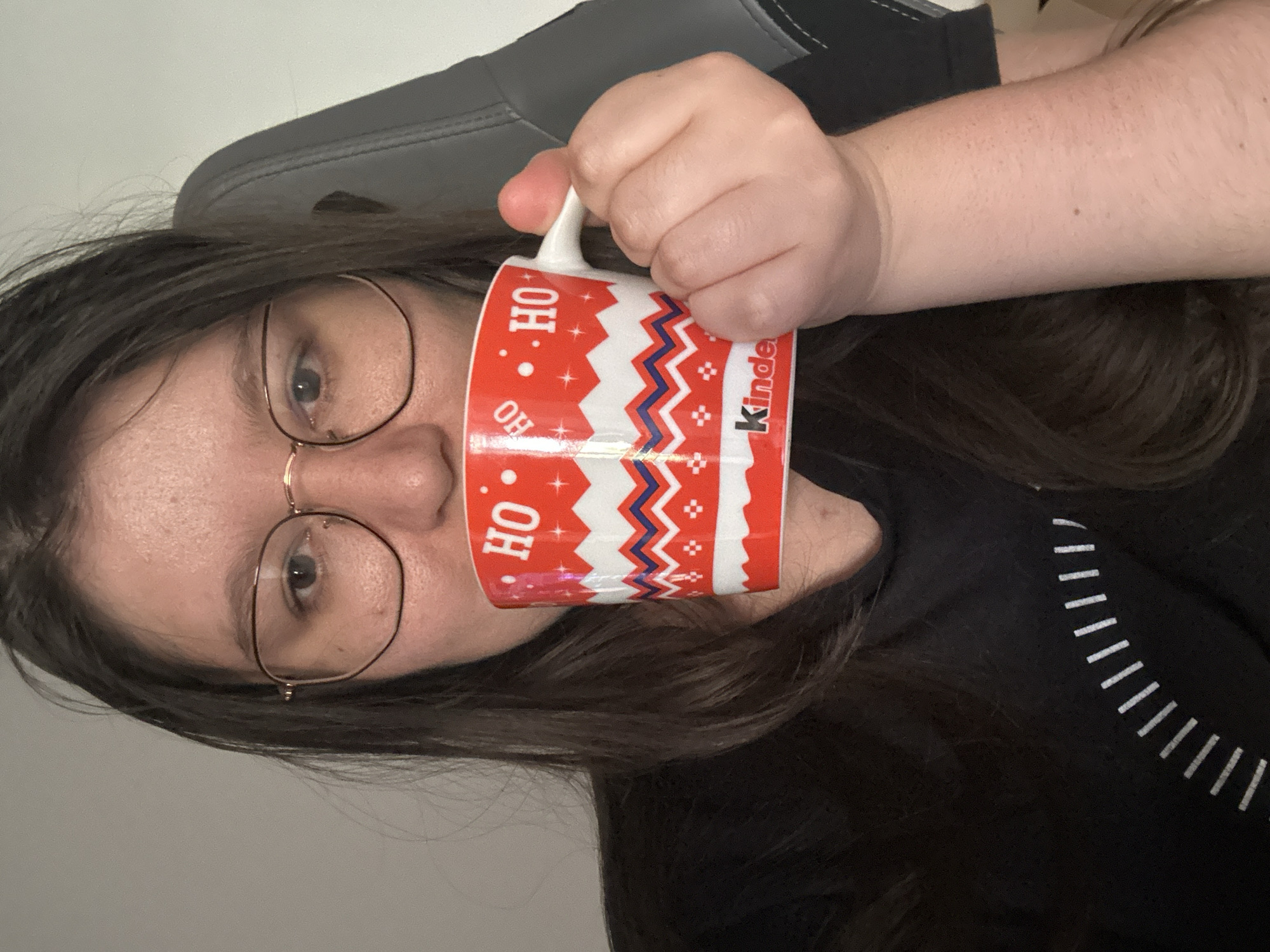 A woman with dark hair and glasses is holding a red and white mug with "OH HO" and "Kinder" printed on it, obscuring part of her face. She is sitting in a gray and black chair. She is wearing a black t-shirt with white dashes. The setting is indoors. The location is not discernible from the image.