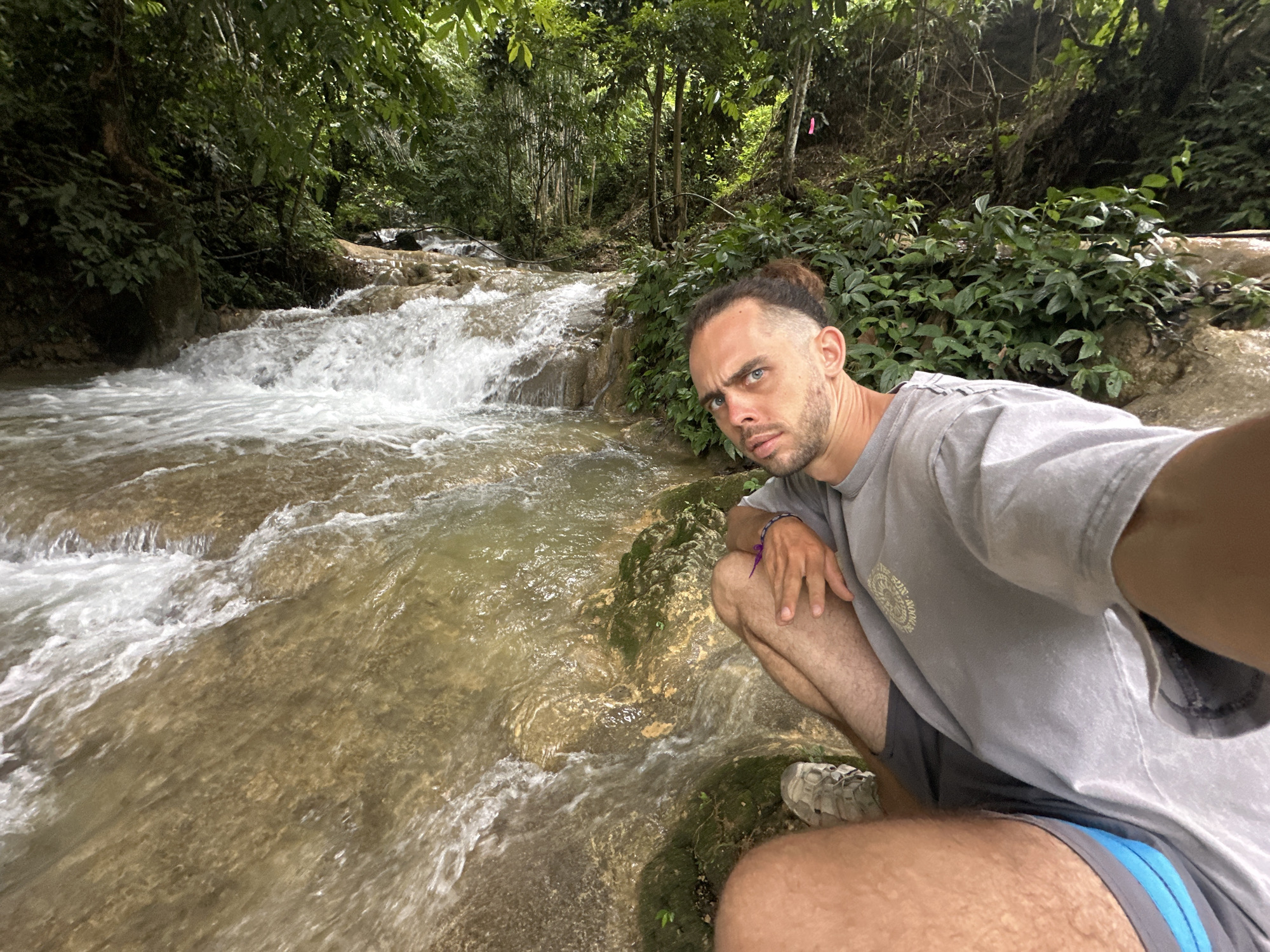 A man is crouching by a rocky stream in a lush green forest. The water is clear and appears to be shallow, with rocks visible beneath the surface. The man is wearing a gray t-shirt and shorts, and he is looking directly at the camera with a serious expression. His hair is pulled back in a bun, and he has a beard. The forest is dense with trees and foliage, and sunlight filters through the leaves, creating a dappled effect. A pink ribbon is tied to a tree in the background, suggesting this may be a marked trail or a point of interest. The scene appears to be in a natural, outdoor setting, possibly a park or a hiking trail.