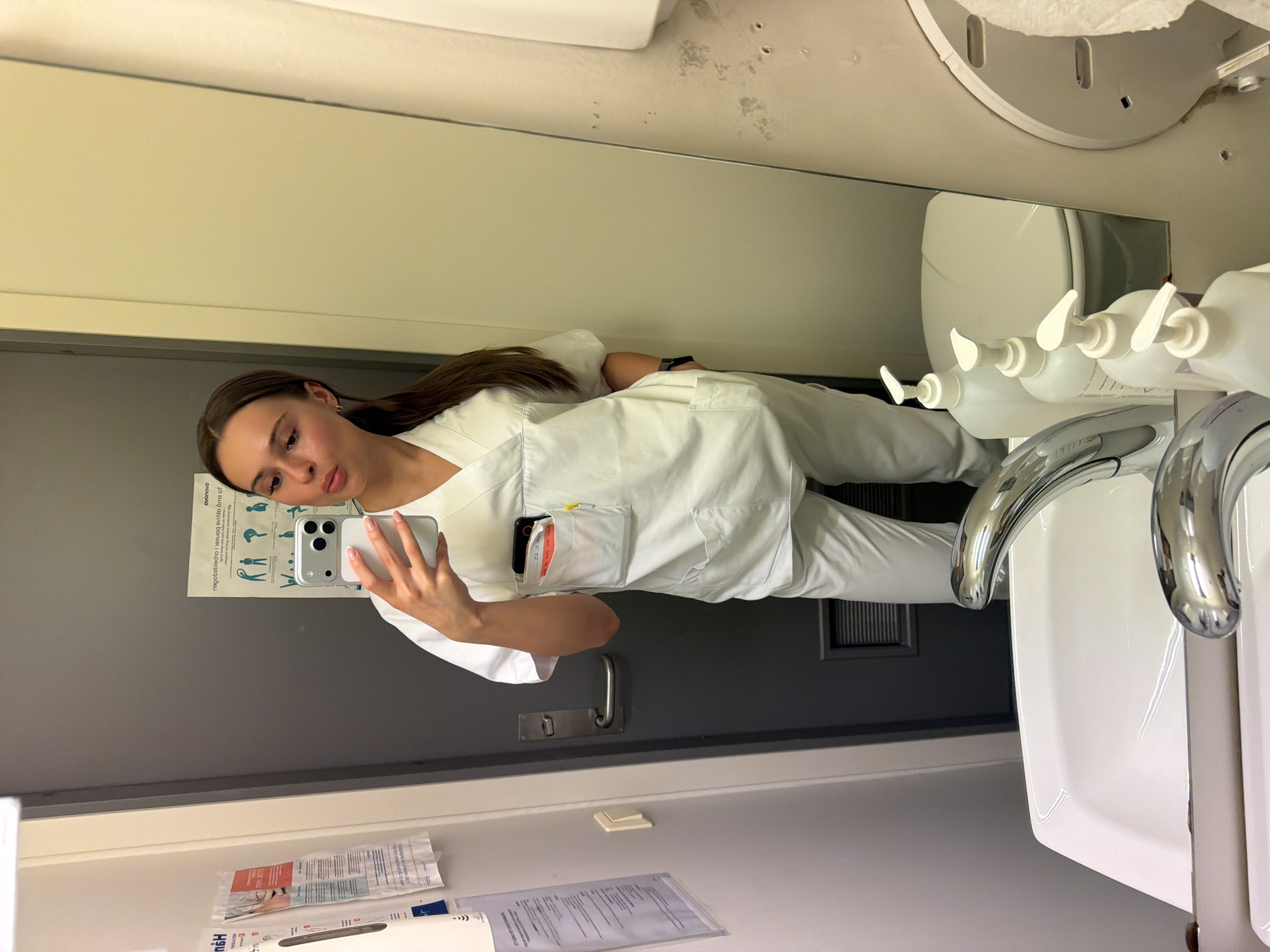 A young woman with long brown hair, wearing white scrubs and holding a smartphone, takes a mirror selfie in what appears to be a public restroom. The background shows a gray door with a silver handle, a white wall, and a sink with chrome faucets. In the reflection, a toilet and several pump bottles of liquid soap are visible. The woman's attire suggests she may be a healthcare professional. The scene is sterile and functional, indicative of a medical or public facility. Harstad, Norway is a city in Northern Norway known for its stunning natural beauty and rich maritime history.