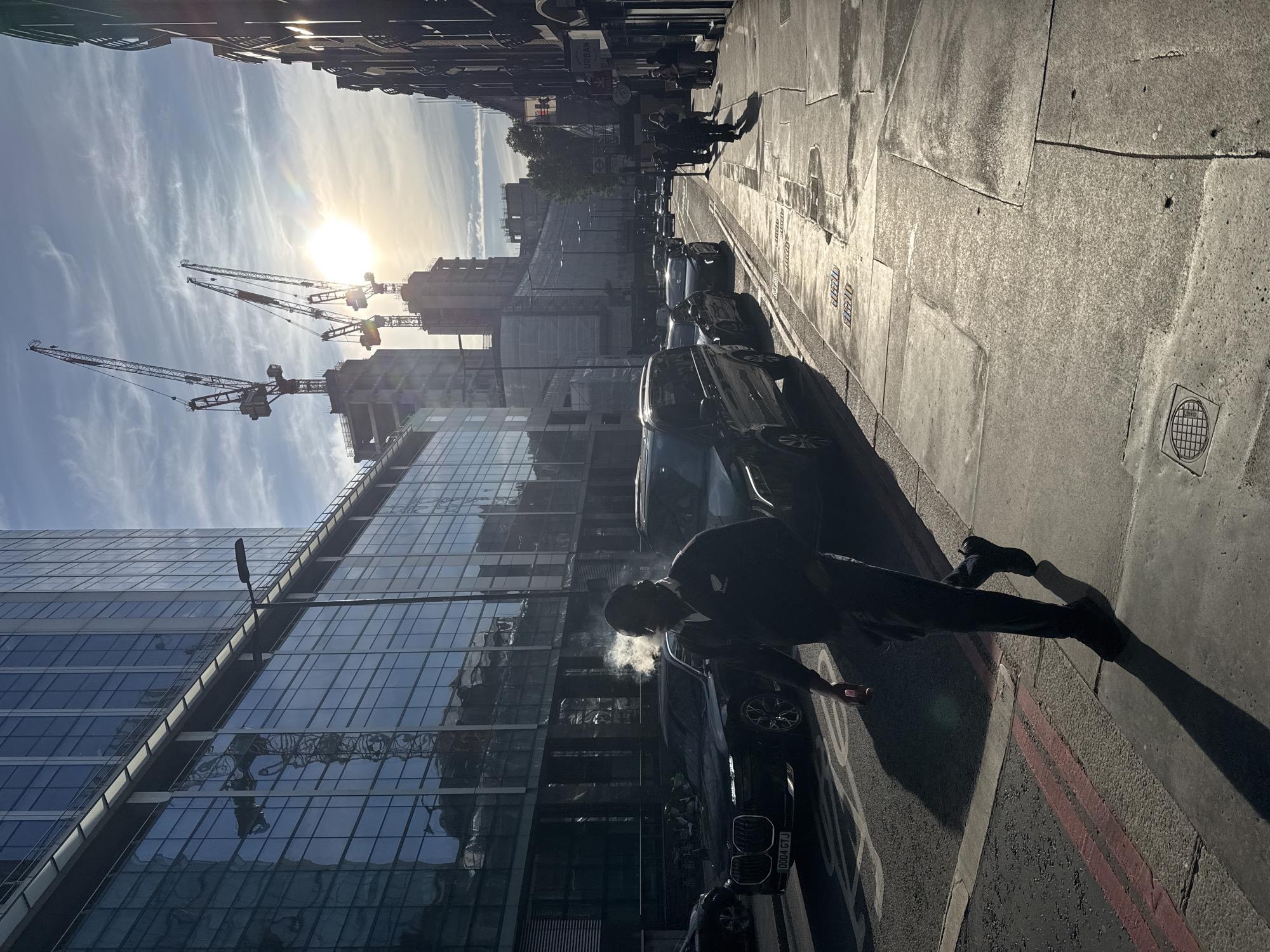 The image depicts a street scene in a city, likely in Europe based on architecture and signage. The viewpoint is oriented vertically, looking down the street.
Foreground: A person is standing beside a parked car, exhaling what appears to be vapor (possibly from a cigarette or vape).
Midground: Parked cars line the right side of the street. Further down, pedestrians are walking along the sidewalk. The street surface appears to be asphalt with lane markings.
Background: Modern glass-clad buildings rise on the left side, reflecting the sky. Cranes are visible in the distance, indicating ongoing construction. The sky is bright with a sunburst effect.
City details: A sign labeled "URBAN" and a tube station logo are visible, suggesting this might be London. The license plate of the vehicle near the smoking person is partially visible, showing "0004 GTJ".
