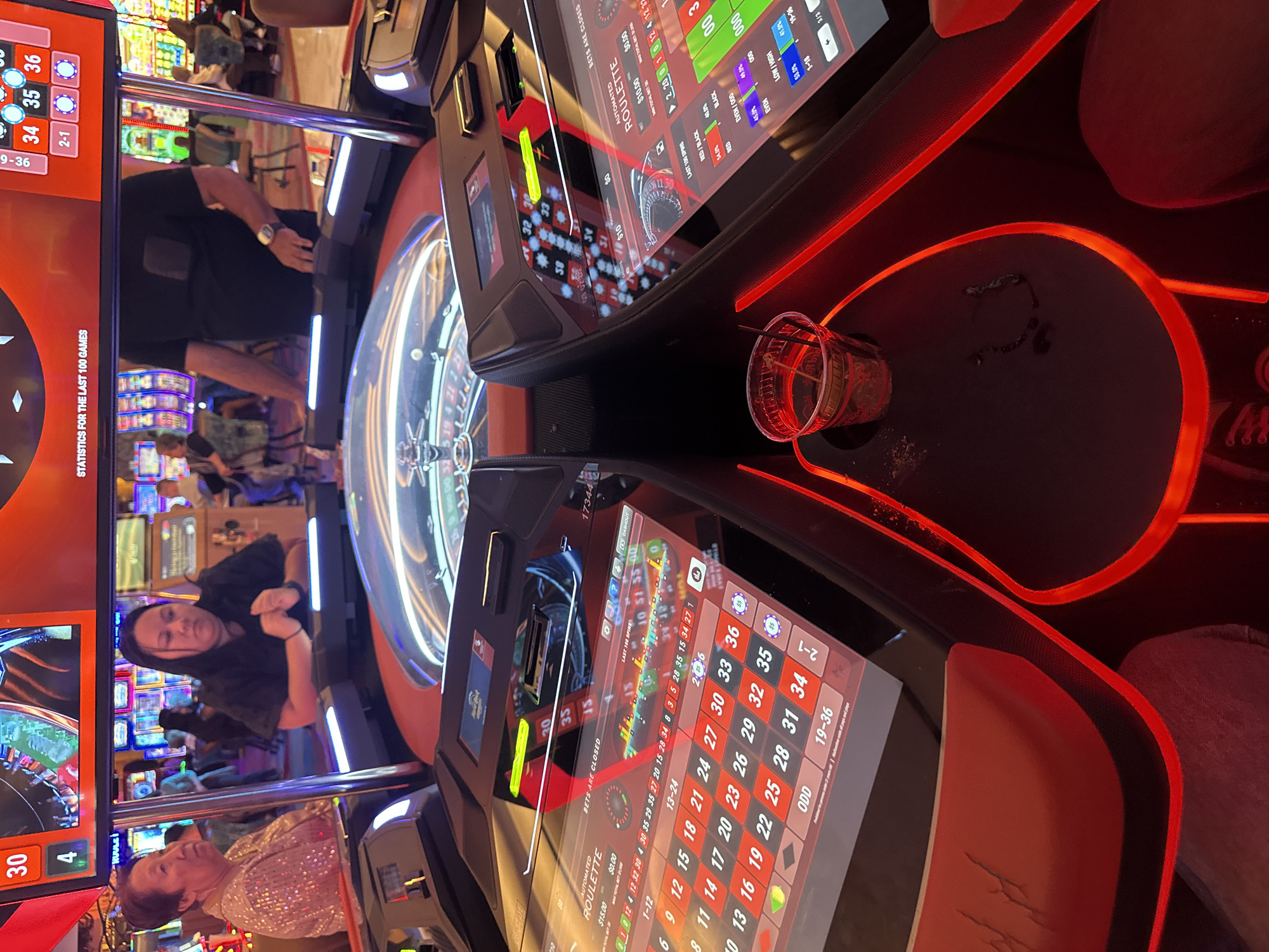 This image depicts the interior of a casino on the Hollywood Seminole Reservation in the United States. The primary focus is an electronic roulette table, featuring a central roulette wheel surrounded by individual player consoles. Each console has a touchscreen display showing betting options and game information. Red LED lighting illuminates the table's edges and features. A partially consumed drink sits on the table. In the background, other patrons are seen at various gaming machines. The lighting suggests a nighttime or indoor environment. The scene conveys a casual atmosphere with people engaged in gambling activities.