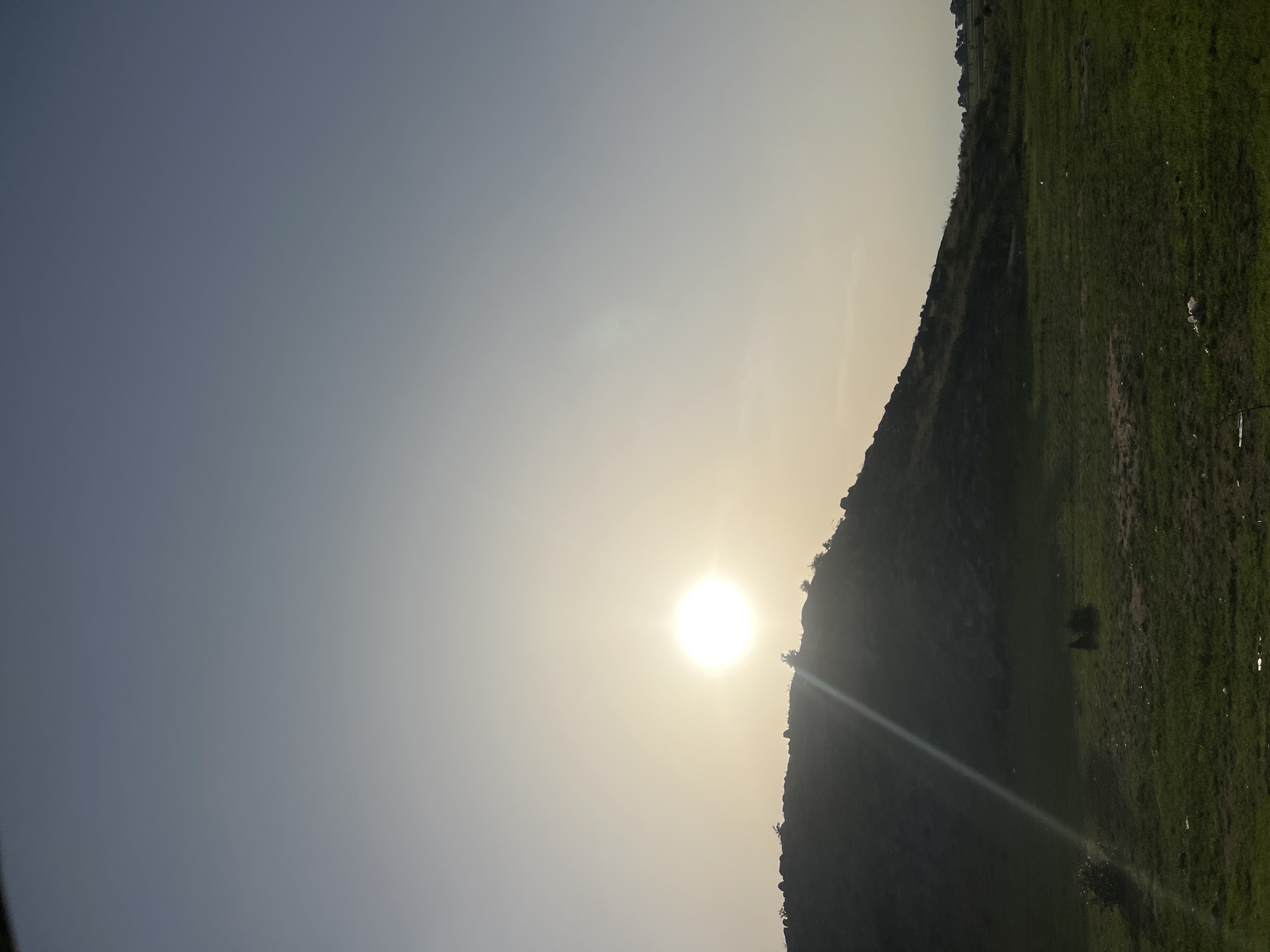 The image captures an outdoor landscape at either sunrise or sunset, situated within or near the city of Bauchi, Nigeria. A brilliant, circular sun is prominently positioned on the upper left, emitting a distinct, diagonal lens flare extending downwards across the scene. The sky exhibits a gradient, transitioning from a luminous, hazy yellow-white around the sun to a desaturated blue-grey in the upper and right sections. A dark, silhouetted hill, characterized by rocky formations and sparse vegetation along its crest, spans the midground from the left to the center-right. The foreground consists of a field of green grass mixed with patches of exposed, drier earth. Scattered white and dark fragments, identified as litter, are visible across the grassy areas in the lower portion of the frame. No individuals, animals, or specific actions are depicted.