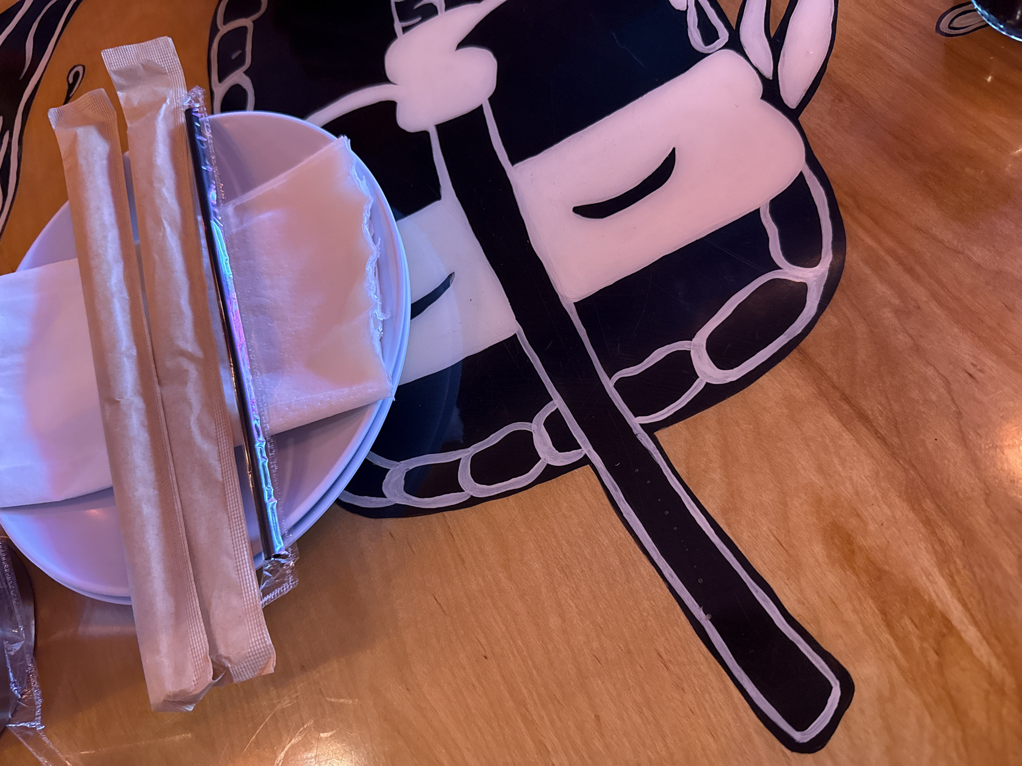 A high-angle, medium shot depicts a setting at a venue in Hollywood, USA. The scene features a small white plate containing paper napkins and two beige, sealed paper straws. A black, plastic-wrapped straw is also present within the plate. Next to the plate sits a black and white placemat with a stylized design of a face. The placemat is positioned on a wooden table with a light-brown grain.