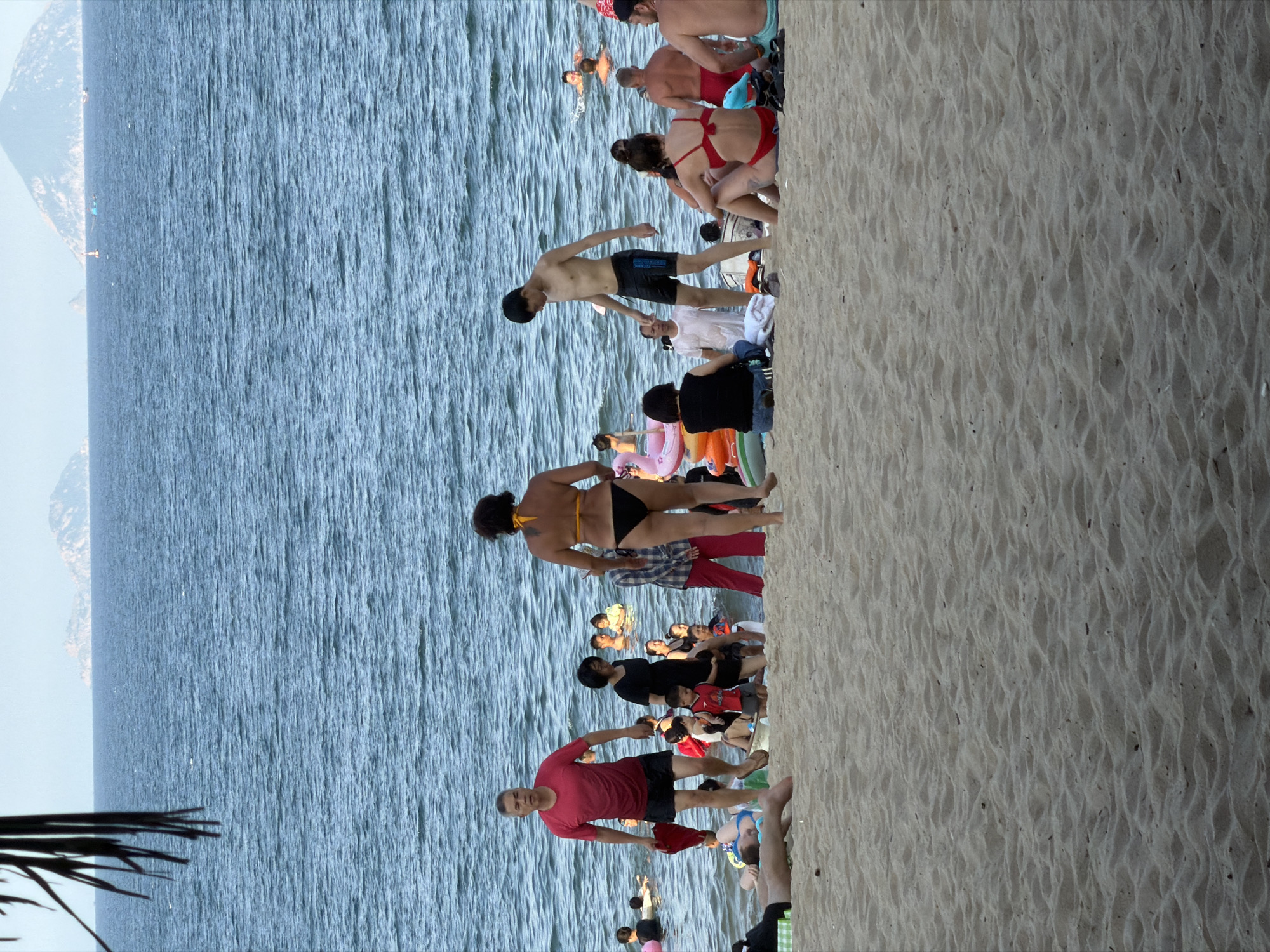 The image depicts a crowded beach scene in Nha Trang, Vietnam. The foreground is dominated by rippled sand, indicative of a beach environment. In the mid-ground, a variety of people are present. Some are in the water, while others are on the shore. Several individuals are seen wearing swimwear, and some are engaged in activities like swimming or sitting on the sand.
In the water, people are scattered, with some swimming and others possibly wading. Several inflatable pool toys, including a pink flamingo and orange rings, are visible, suggesting recreational use of the water.
On the beach, there is a mix of people sitting, standing, and walking. A man in a red t-shirt and black shorts is walking towards the water, holding a red item. A woman in a black bikini is walking away from the camera. Another woman in a black dress is standing near a child in a red shirt. A young man in black swim shorts is standing and looking down. A group of people are sitting on the sand, some in swimwear.
In the background, the ocean stretches out to the horizon, with gentle waves. In the far distance, islands or coastal mountains are visible, with a hazy sky above. The overall impression is a lively and populated beach on a clear day.
