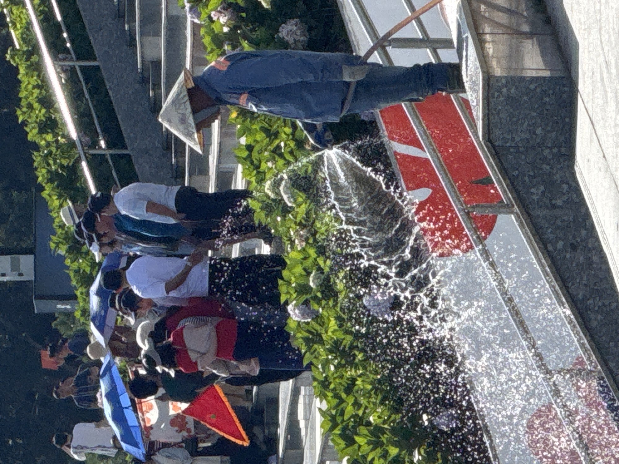 The image captures a street scene in Da Lat, Vietnam, likely near a public area with steps and greenery. In the foreground, a person wearing a conical hat and blue work clothes is watering plants with a hose. The water spray creates a shimmering effect. In the mid-ground, a crowd of people are gathered, some holding colorful umbrellas and a red flag. The background shows lush green foliage and a portion of a building or structure. The overall impression is of a warm, sunny day, with a mix of tourists and locals in a vibrant outdoor setting.