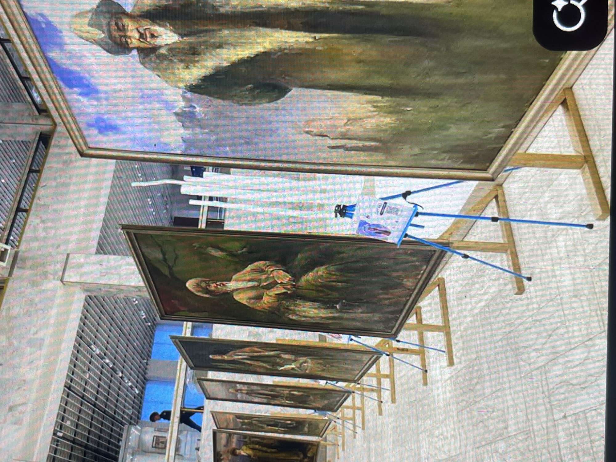 The image depicts an indoor exhibition hall, likely a museum or gallery, in Bishkek, Kyrgyzstan. The hall features a modern architectural design with a suspended ceiling grid, lighting fixtures, and large windows or open spaces that reveal a blue sky or interior lighting. Several large, framed oil paintings are displayed prominently on wooden easels arranged in a row. These paintings appear to be portraits of men in traditional attire, set against natural landscapes or plain backgrounds. The style of the paintings suggests a historical or cultural theme. In the foreground, a blue tripod stand supports an information display with a QR code and text, likely providing details about the artworks or exhibition. The floor is tiled with a geometric pattern. In the background, partially visible through an opening or upper level, a person is seen walking. The overall impression is of a well-lit, organized art exhibition space.