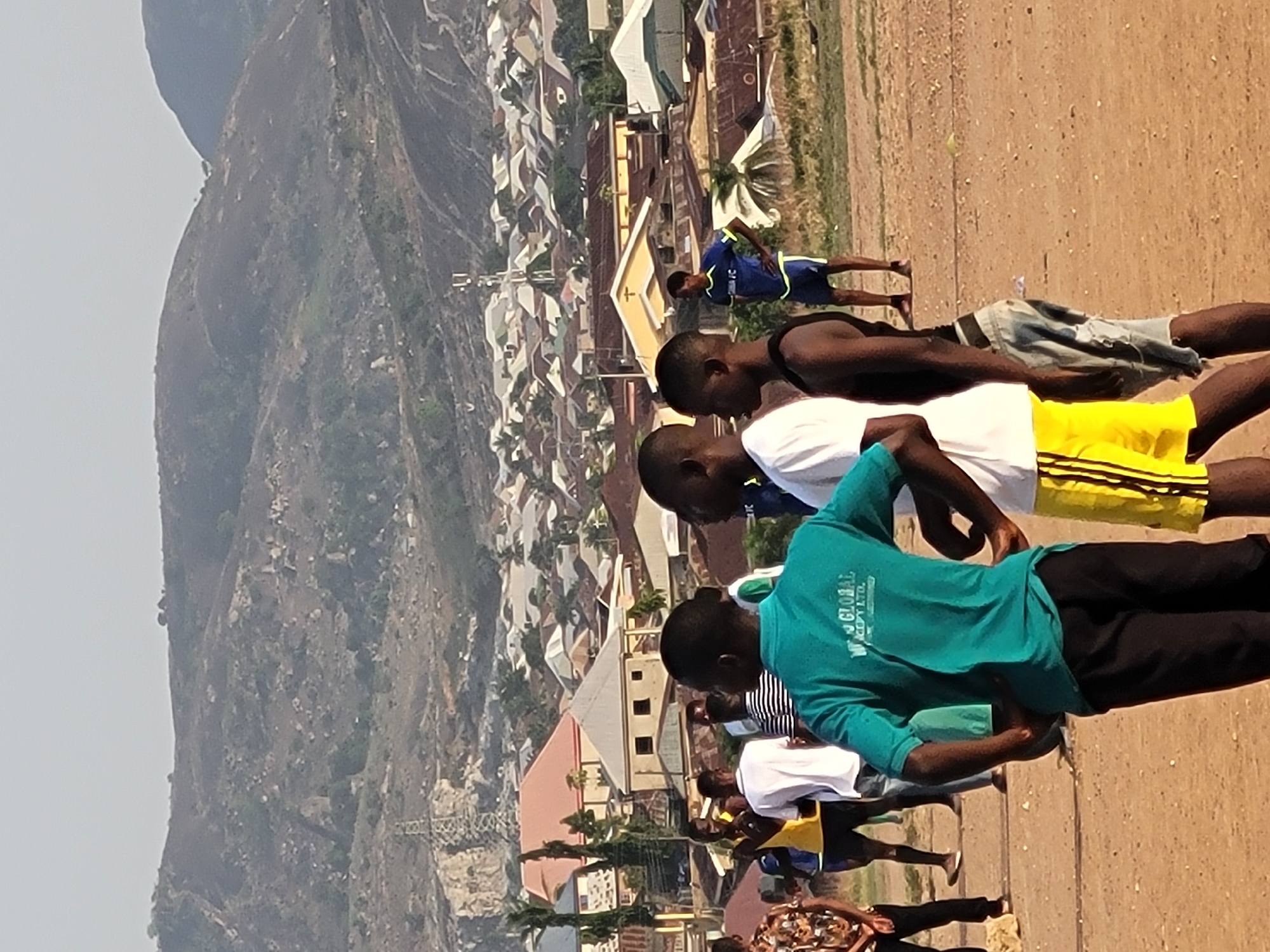 The image depicts a group of people standing on a dirt field in Ibo, Nigeria. In the background, there is a hillside and buildings. The people are wearing a variety of clothing, including t-shirts and shorts. Some of them appear to be exercising or stretching. The overall scene suggests an outdoor activity or gathering in a residential area near a natural landscape feature.