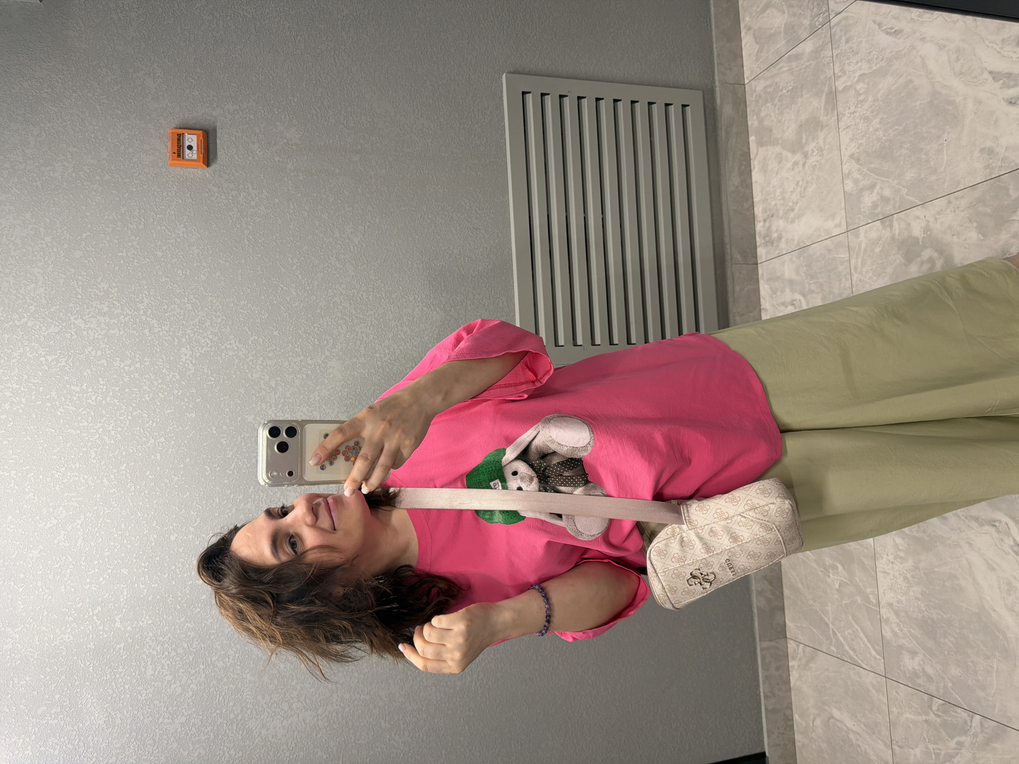 A woman with dark brown, wavy hair is taking a selfie in a room with textured gray walls. She is wearing a bright pink t-shirt with a graphic of a bunny wearing a green cap on the front and light green wide-leg pants. A beige purse with the "GUESS" logo is slung across her body. She is also wearing a bracelet on her left wrist. On the wall behind her, there is a red fire alarm box and a horizontal vent. The floor is tiled with a marble pattern. The scene appears to be indoors, possibly in a public building. There are no specific elements in the image that suggest the location is the city of Perm, Russia.