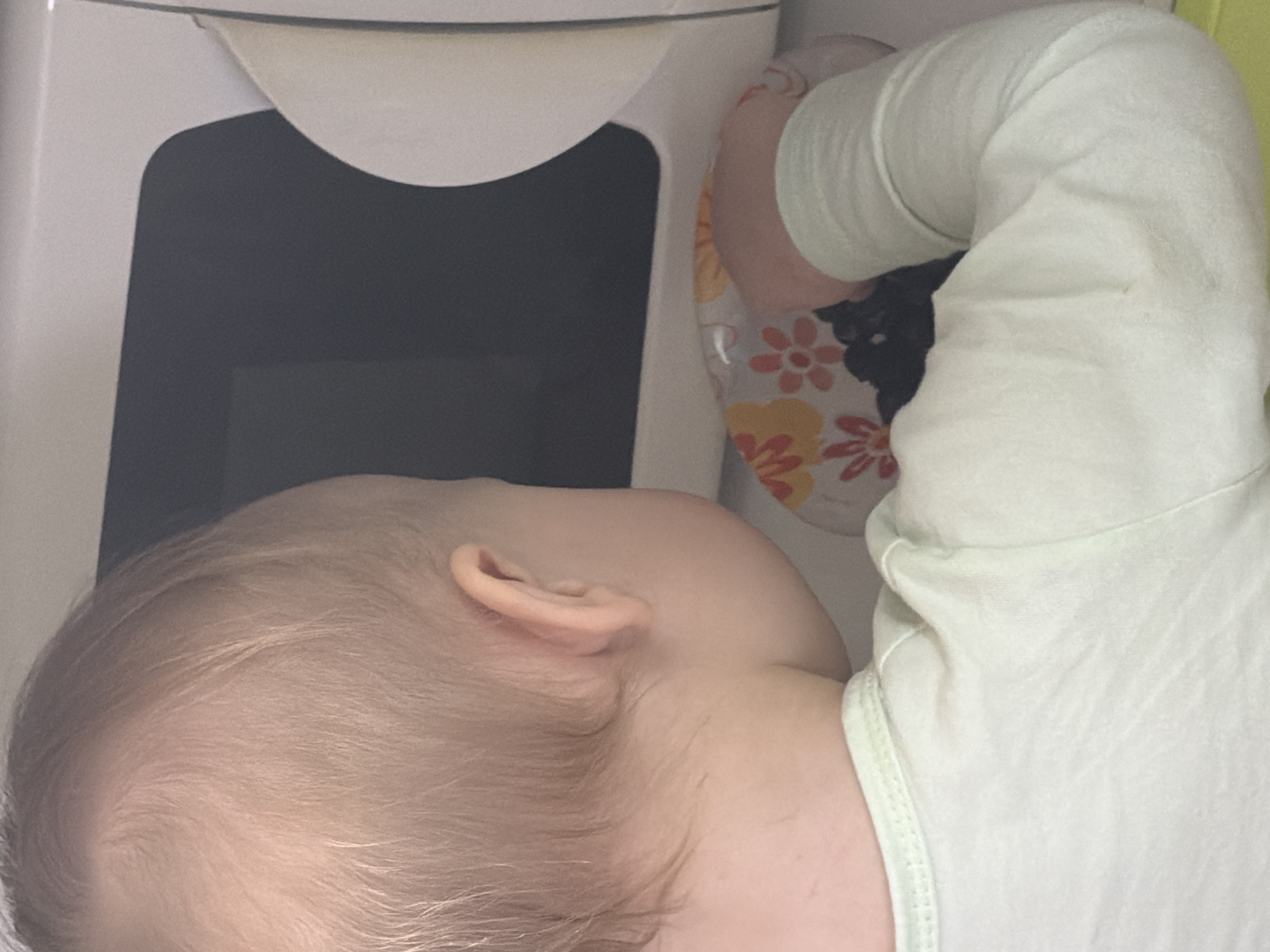 A young child with blonde hair, wearing a light green long-sleeved shirt, is lying down. The child's head is near a white appliance with a dark rectangular screen, possibly a microwave or refrigerator. A floral-patterned fabric is near the child's arm. The location is not discernible.