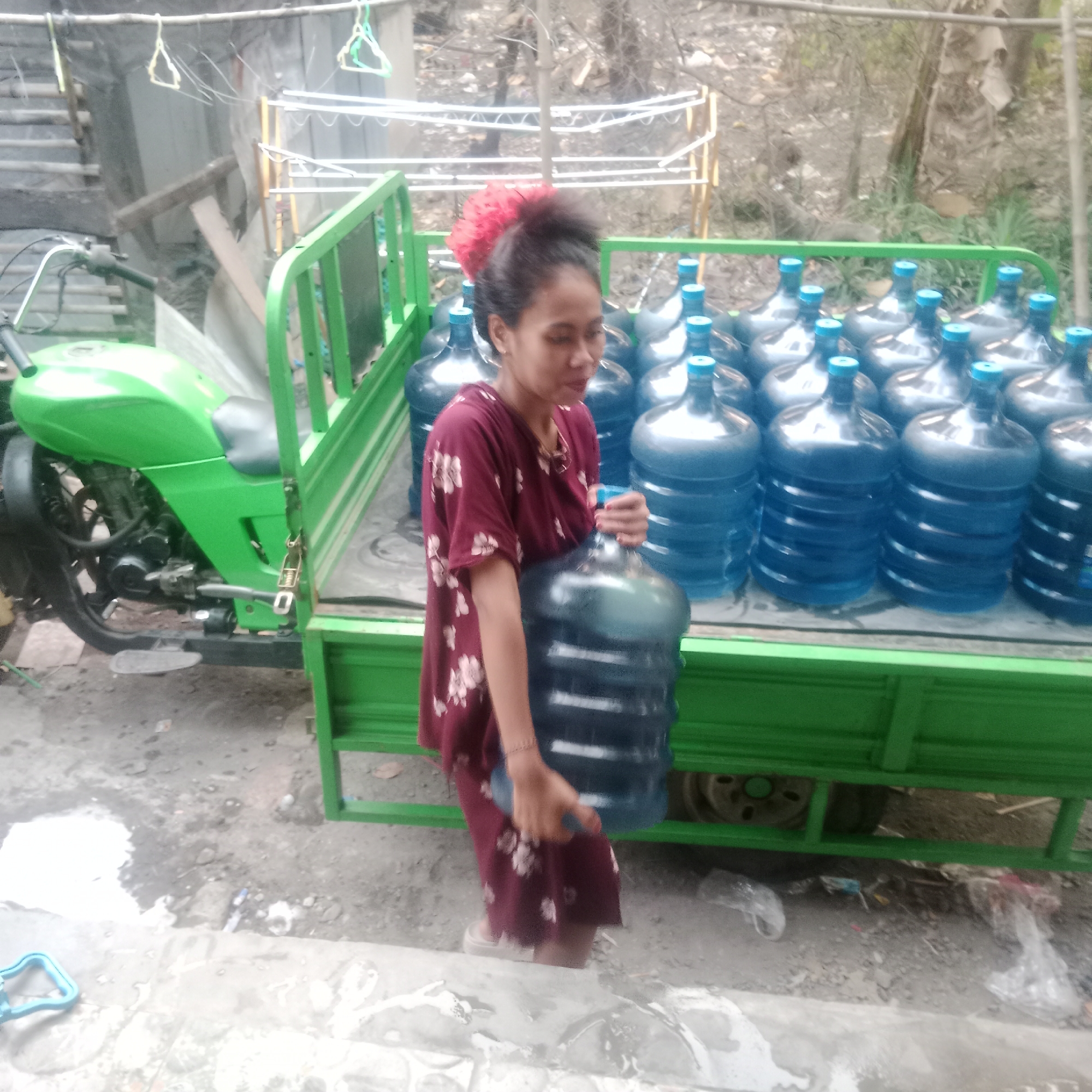In Tegowanu Kulon, Indonesia, a young woman with a bright red hair accessory is seen in the back of a bright green utility vehicle. She is holding a large, clear plastic water jug by its handle, its blue cap visible. The truck bed is filled with numerous similar water jugs, many of them filled with clear liquid, and stacked in rows. A green motorcycle is parked on the left side of the truck, partially visible. The ground beneath the truck is a mix of dirt and concrete, with some scattered debris. In the background, a makeshift clothesline with hangers and some laundry is visible, along with trees and other structures.