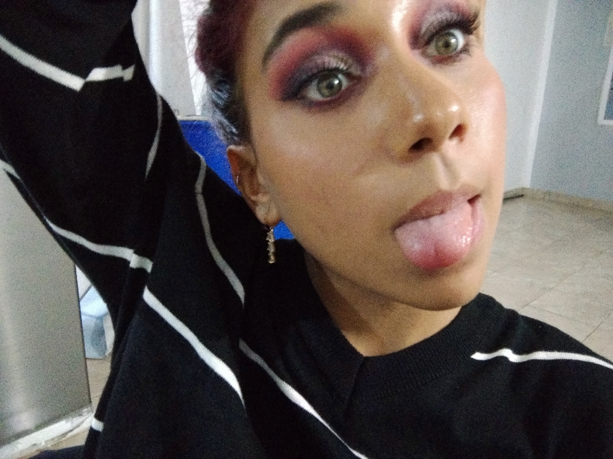 A close-up, slightly blurry, over-the-shoulder selfie shows a young person with maroon-dyed hair and dramatic eye makeup, featuring pink and purple eyeshadow and false eyelashes. The person is sticking their tongue out, which appears to be slightly red. They are wearing a black long-sleeved shirt with white diagonal stripes. A small silver earring is visible. The background is out of focus and shows a light-colored wall and floor, with a blue object to the left. The scene appears to be indoors. There is no discernible location in Parroquia Charallave, Venezuela.