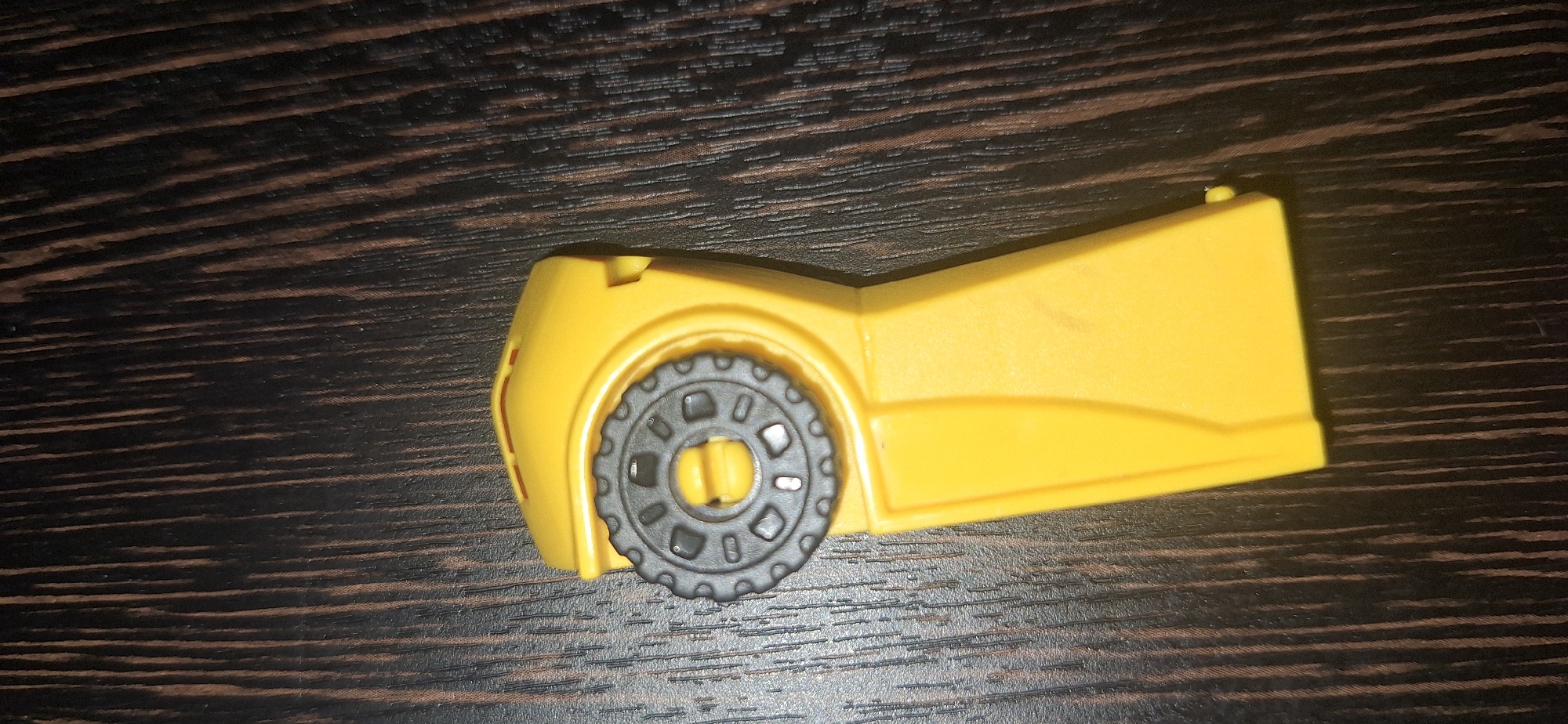 A bright yellow, plastic toy piece is positioned on a dark wood grain surface. The toy has a rounded top section with a gray, circular component featuring a gear-like edge and internal cutouts. A yellow protrusion is visible in the center of the gray component. Below the rounded top, the yellow plastic extends into a longer, tapered section. The wood grain surface appears to be a table or countertop with vertical, dark brown lines. The image is a close-up, top-down view of the toy. There is no information about its location in Bila Tserkva, Ukraine.