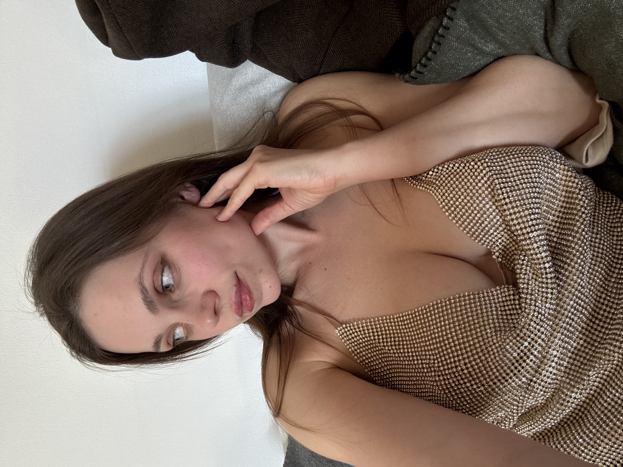 A woman with long brown hair is lying on a bed or soft surface. She is wearing a sparkly gold top. Her right hand is near her face, touching her cheek. The background includes a white wall and a brown blanket or coat. The scene's location in Sappada, Italy cannot be determined from the image.