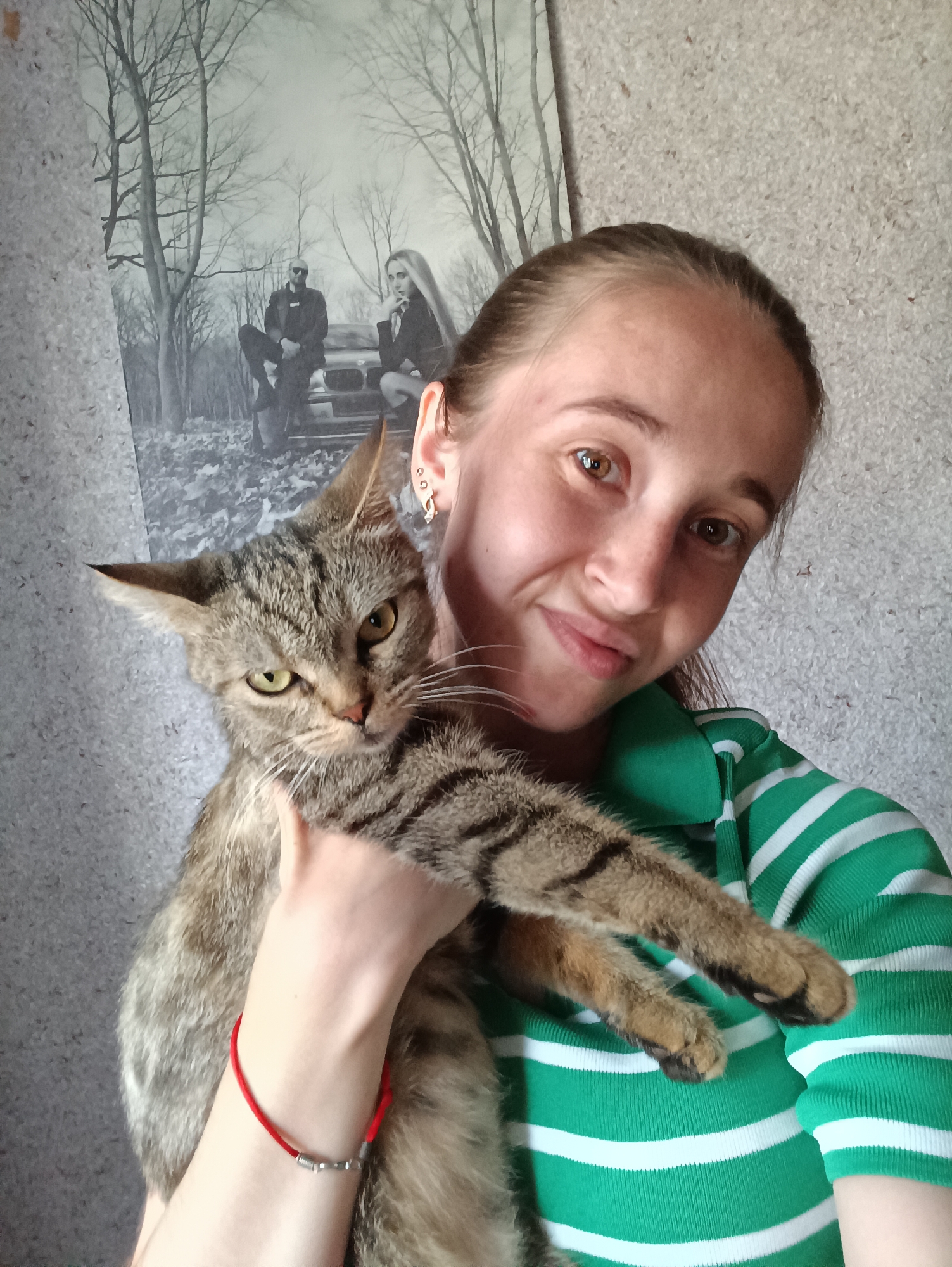 A young woman is holding a tabby cat in her arms, posing for a selfie. The woman is wearing a green and white striped shirt. The cat is looking directly at the camera with a slightly annoyed expression. In the background, there is a framed black and white picture of a man and a woman sitting in front of a car in a wooded area. The wall behind them appears to be textured. The location is Vypasne, Ukraine.