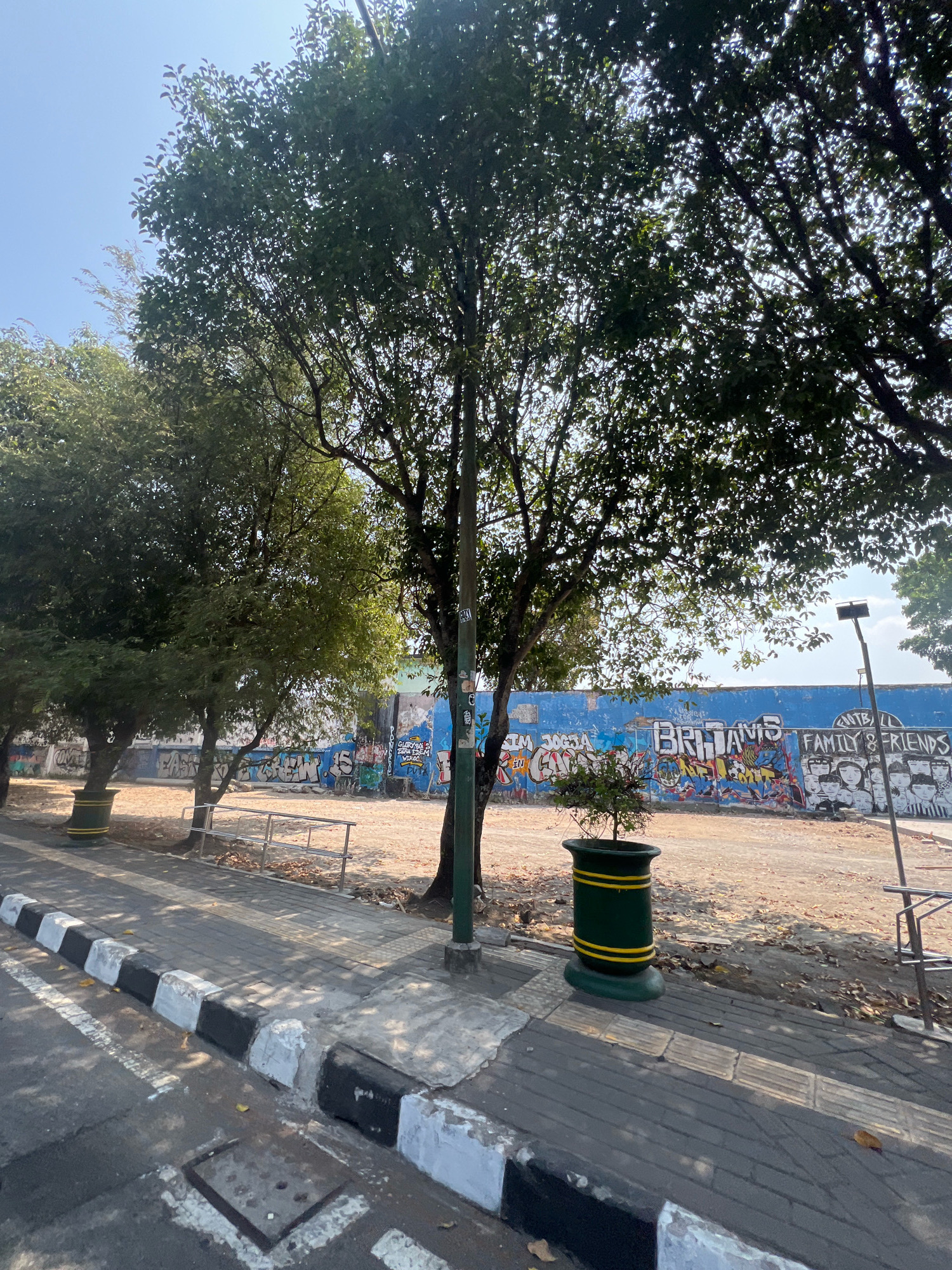 The image captures a sun-drenched scene in Yogyakarta, Indonesia, likely near the Tembok Sebelah Barat, a popular graffiti art location. The foreground features a sidewalk with a distinctive black and white painted curb, leading to a street. Potted plants with green and yellow accents are placed along the sidewalk. In the mid-ground, a metal railing separates the sidewalk from a sandy, open area. The background is dominated by a vibrant blue wall covered in a variety of graffiti tags and murals. Notably, one mural depicts a group of figures, and another spells out "BRITAMIS." Trees with lush green foliage are scattered throughout the scene, casting dappled shadows. The sky is a clear, bright blue, indicating a sunny day. The overall impression is an urban outdoor space adorned with street art and natural elements.