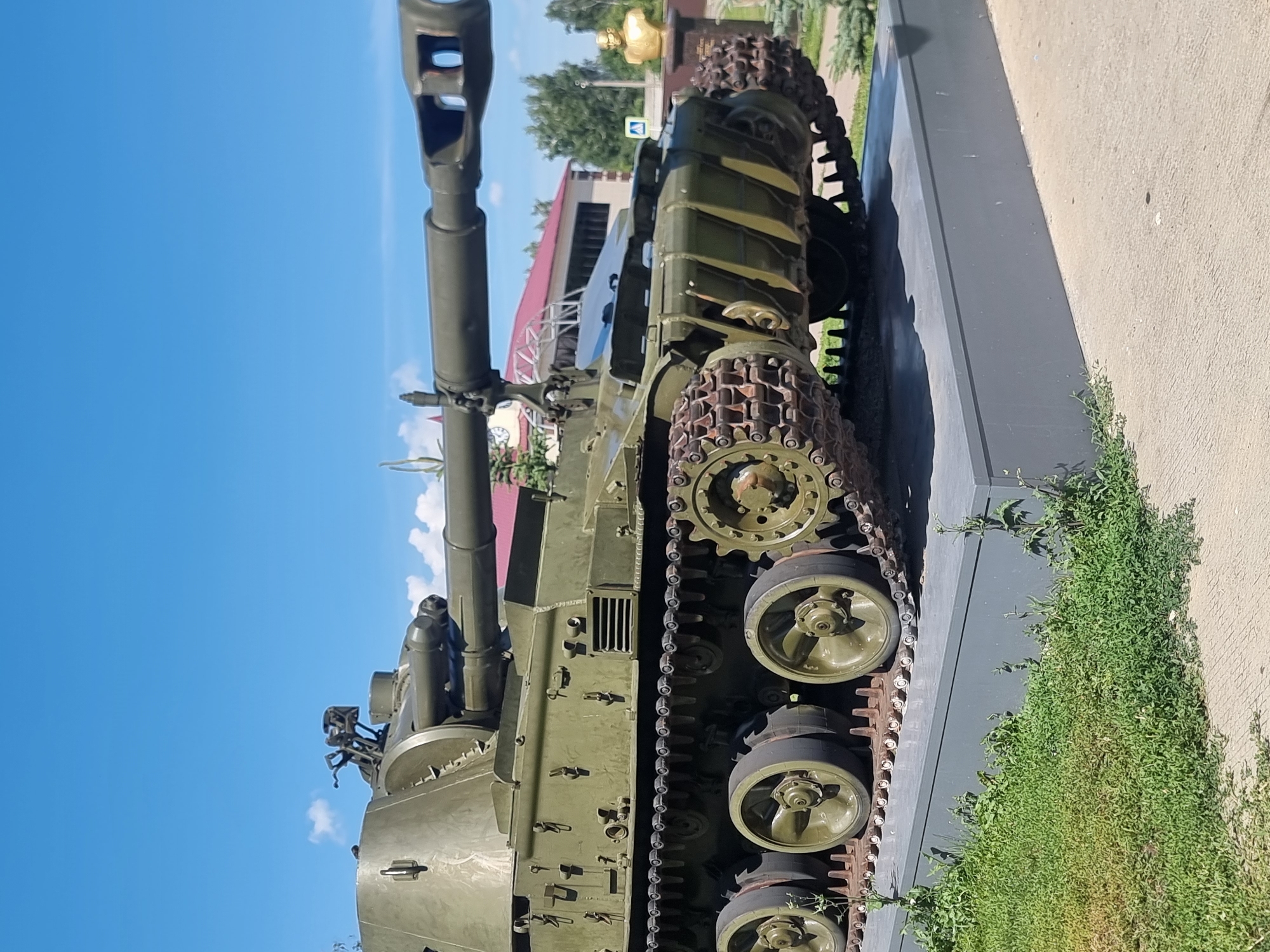 The image shows a view of a green self-propelled gun, likely a memorial, displayed on a concrete pedestal. The vehicle is positioned outdoors under a clear blue sky with some white clouds. The gun barrel is prominent, extending horizontally. The track system and road wheels of the vehicle are clearly visible. In the background, there are buildings with red roofs, trees, and a golden bust on a dark pedestal. The ground in front of the pedestal is paved, and there is green grass and some foliage around the base. The image is taken from a low angle, looking up at the self-propelled gun.