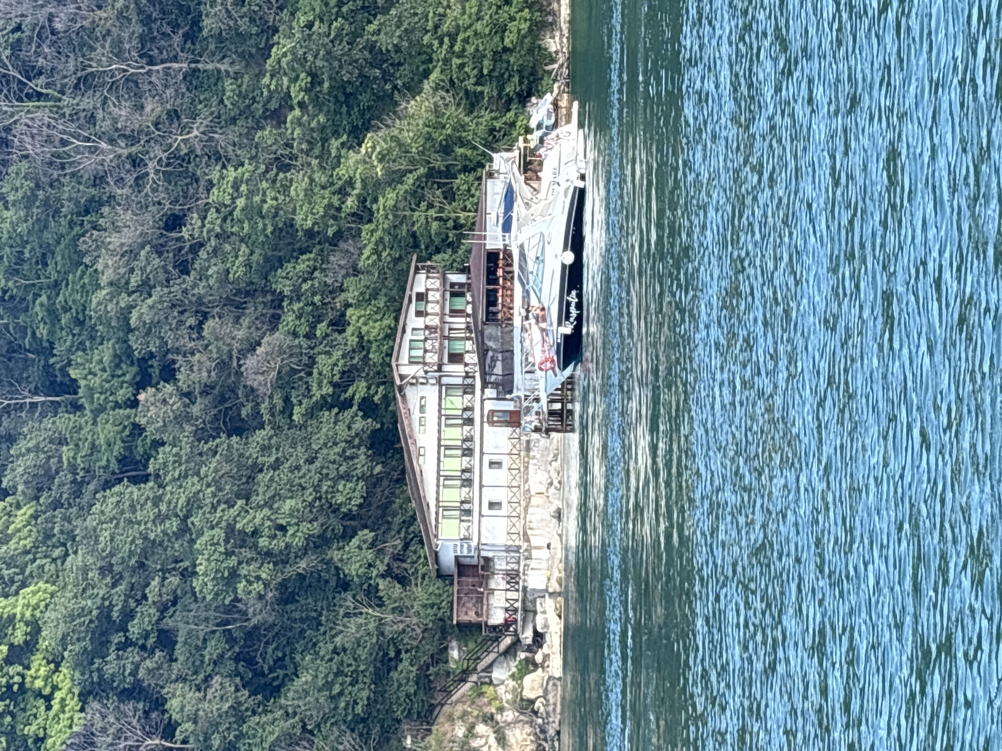 This is a medium-angle, eye-level, vertical shot of a building and boats on a body of water, with a densely wooded hillside in the background. The building, a multi-story structure with white walls and a brown roof, is situated on a rocky incline. It features numerous windows, balconies with wooden railings, and some green awnings. A wooden staircase leads up the side of the building from the water's edge. In the foreground, two boats are docked. The larger boat, a white motor yacht with a dark hull, has the name "Rasputin" visible on its side. The water in the foreground is choppy and a vibrant turquoise color. The background is dominated by a steep hillside covered in lush green trees. The scene appears to be a coastal or lakeside resort.