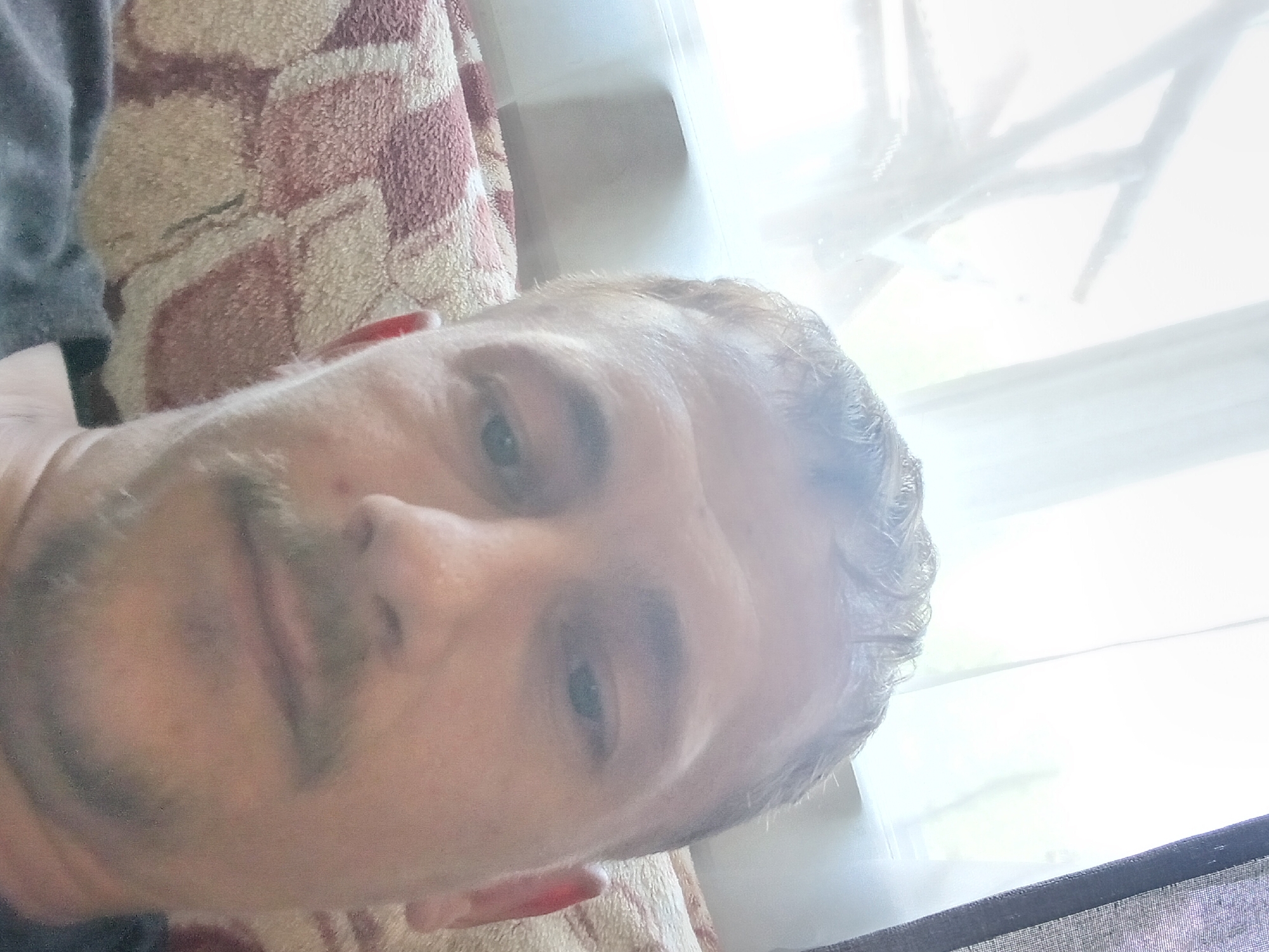 The image is a close-up, slightly blurry, selfie of a man in Verkhniachka, Ukraine. He is lying down on a patterned couch with a pink and beige design, covered in a blanket with similar colors. The man has short, light brown hair, a thin mustache, and a light beard. He is looking directly at the camera, which is positioned above him. Behind him, a window is visible with sheer white curtains and some blurry green foliage outside, suggesting it is daytime. To the right of the window, a dark grey textured curtain is partially visible. The overall lighting is bright, with some glare from the window.