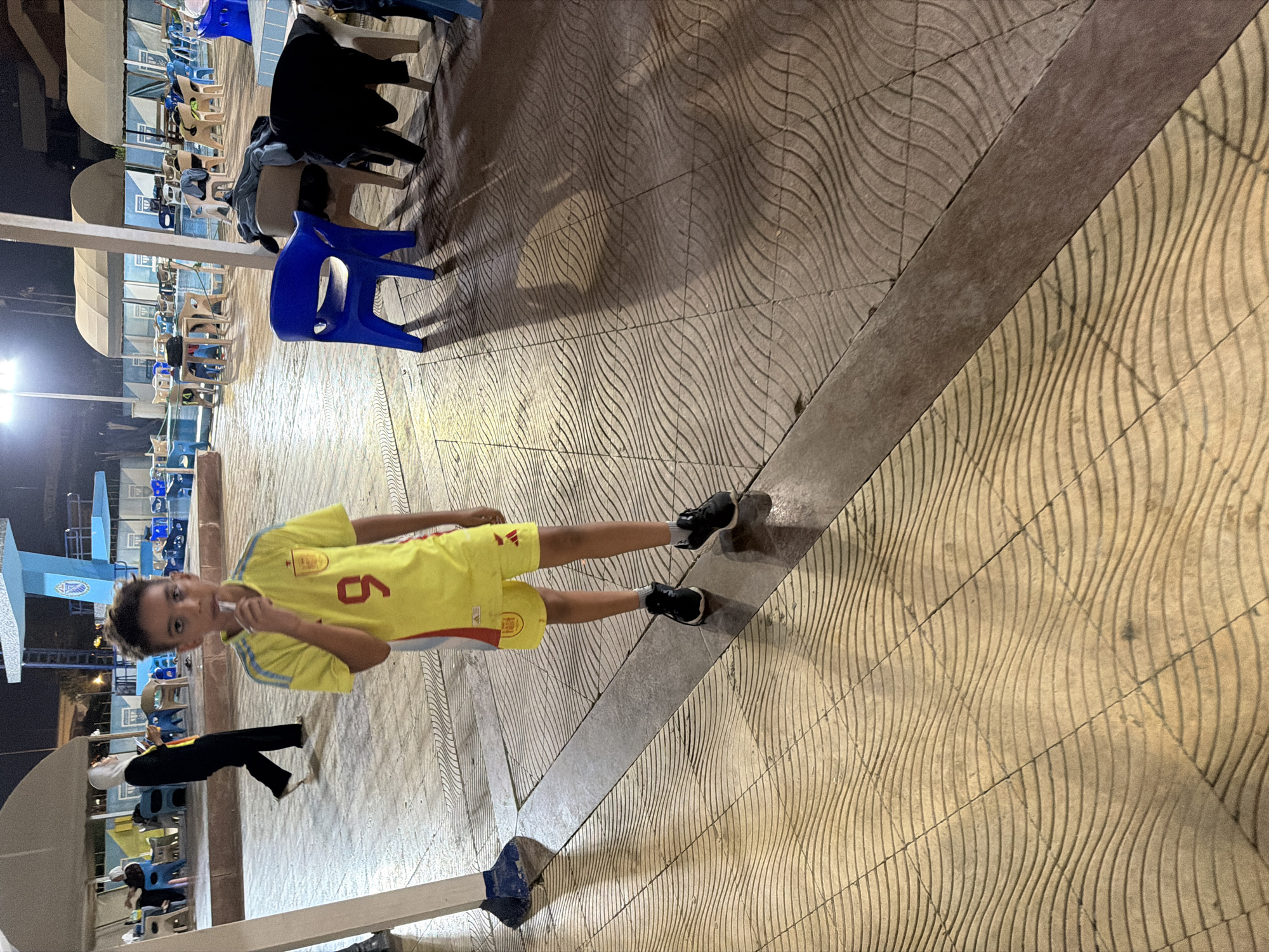 A young boy stands in a courtyard at night, holding a lollipop to his mouth. He is wearing a yellow soccer jersey with red and blue accents, and matching yellow shorts. He has black athletic shoes and gray socks. The boy is the central figure in the image, with a portion of a pool area visible in the background. The ground is paved with patterned tiles, and shadows from overhead structures and lighting are cast across the area. In the background, other people are visible near a pool, and there are several blue plastic chairs scattered around. The setting appears to be an outdoor recreational area, likely a pool facility, located in 6th of October City, Egypt, given the context. The lighting suggests it is evening or night.