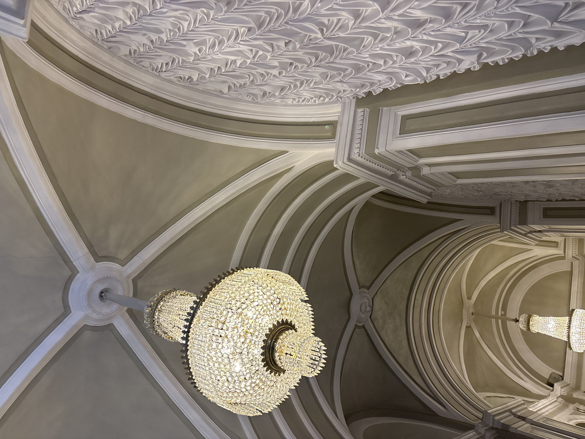 The media file shows a close-up, low-angle shot of the ornate interior of a grand building, likely a palace or museum, in Saint Petersburg, Russia. The primary subjects are two large, elaborate crystal chandeliers hanging from a highly decorated ceiling. The ceiling itself features intricate white molding and recessed paneling, with a distinct ruffled, fabric-like texture in the upper portion of the image. The architectural style suggests a historical European setting.
The chandeliers are the focal point of the image. They are multi-tiered and appear to be made of numerous small crystals, catching and reflecting light. One chandelier is more prominently featured in the foreground, while another is visible in the background. The lighting is soft and warm, emanating from the chandeliers themselves, suggesting the scene might be captured during the evening or in a dimly lit interior.
The setting is characterized by a sense of opulence and grandeur, common in historic Russian architecture. The curved lines of the ceiling and the detailed moldings create a dramatic and elegant atmosphere. There are no people visible in the frame, nor any specific activity or event taking place. The image focuses solely on the architectural details and the decorative elements of the interior. The colors are muted, dominated by shades of beige, cream, and white, which further emphasizes the intricate textures and forms of the ceiling and chandeliers. No text is visible in the media file.
