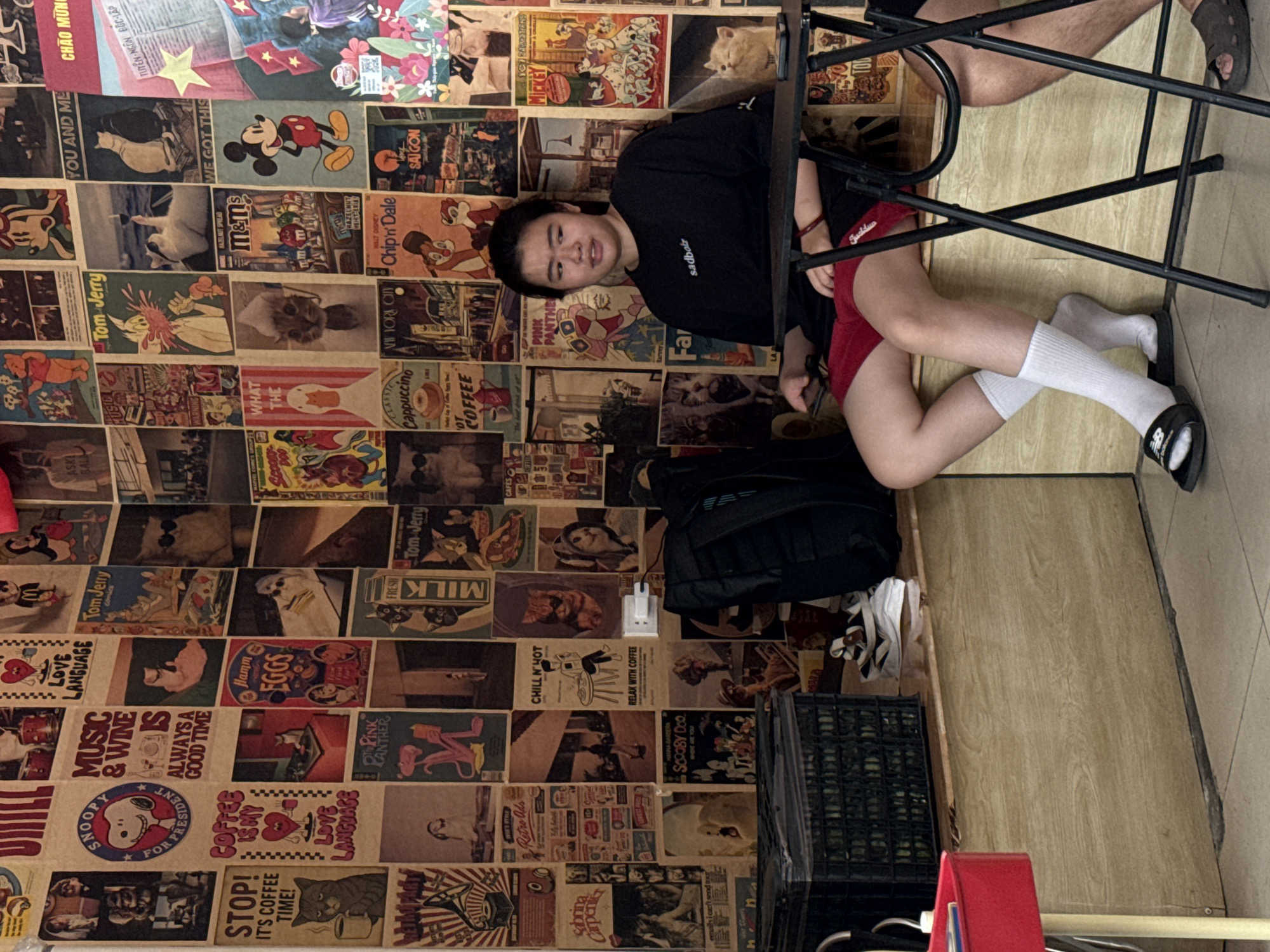 A view of a wall covered in posters, the corner of a room, two people at a table. The poster wall consists of an array of cartoon and food-themed advertisements, including Mickey Mouse, Tom & Jerry, and vintage product labels. A person is sitting with their torso facing the camera, and their legs bent towards the lower right. They wear a black t-shirt with 'sadboiz' printed on it, red shorts, and white socks with black sandals. Another person sits to the upper right, with their legs visible. A black backpack and various storage containers rest against the wall, along with other objects. The floor is wood-colored. This scene is located in Đà Nẵng, Vietnam.