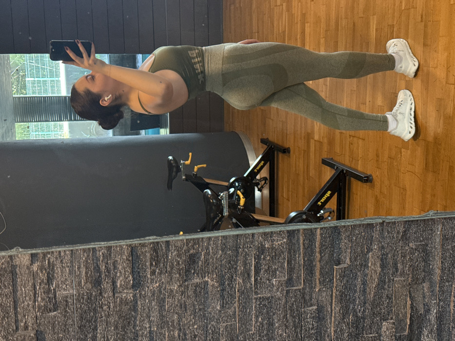 The image displays a female individual taking a mirror selfie within a gym or fitness center, located in Mexico City, Mexico. The subject, positioned with a slight side profile, is reflected in a large mirror. She has dark hair pulled back into a bun, wears white wireless earbuds, and is holding a dark-colored smartphone with her raised left hand, capturing her reflection. She is dressed in an olive green sports bra featuring the word "PINK" and matching high-waisted, textured olive green leggings. Her footwear consists of white athletic shoes with distinctive sole designs and white ankle socks. The immediate environment includes a polished light brown wooden floor and dark grey walls. In the reflected background, two black stationary bicycles with yellow accents are visible. A large window, reflecting outdoor greenery and parts of a distant building, suggests an urban setting beyond the facility. A section of a dark grey stacked stone wall frames the left side of the mirror.