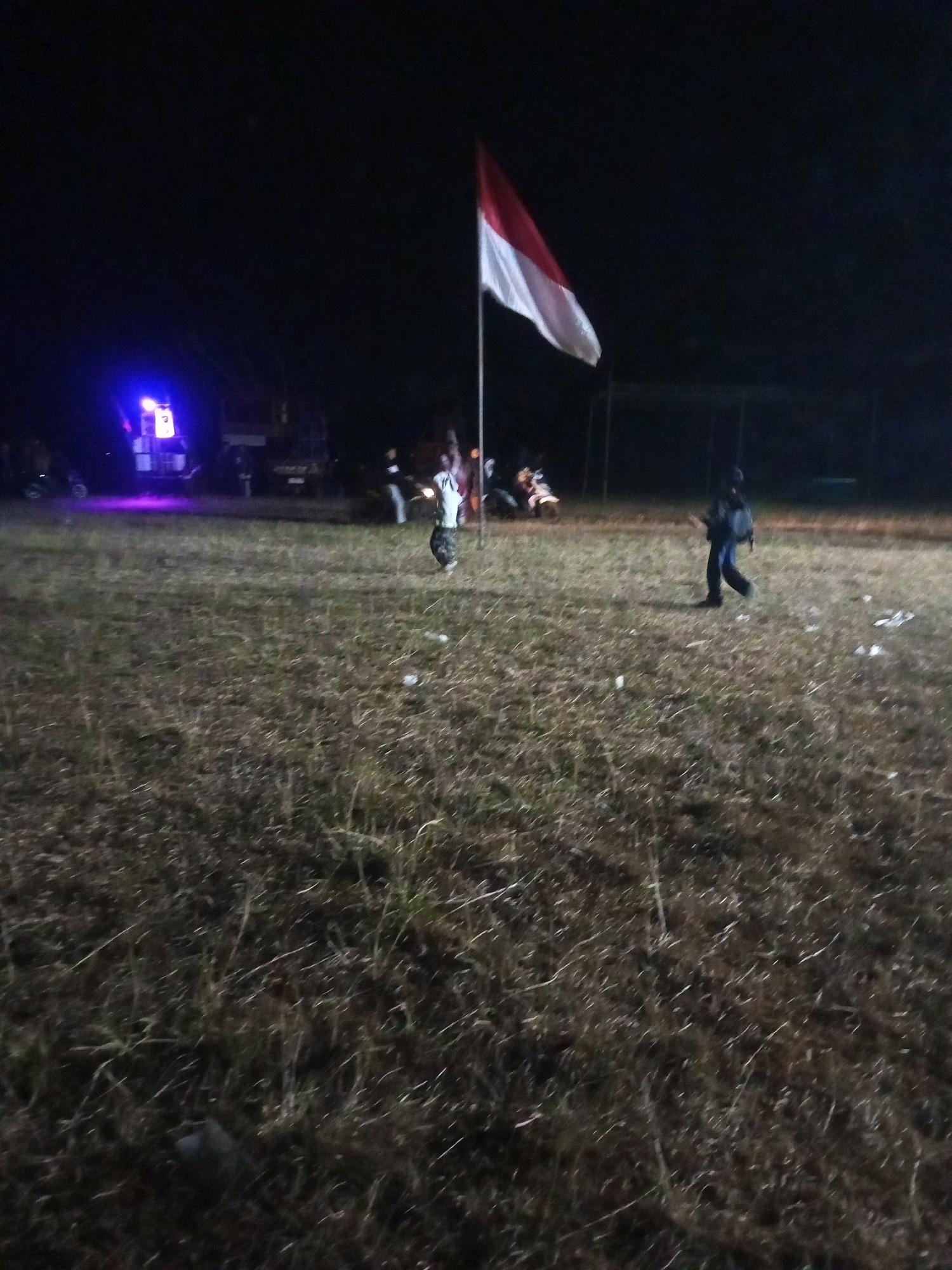 The image depicts an outdoor scene at night, likely in a grassy field, with a prominent Indonesian flag flying from a flagpole. The flag is red and white, indicating its national significance. Several individuals are visible in the scene, some appearing to be in motion. In the background, there are lights from what appears to be a sound system or stage setup, with a blue glow emanating from it. Vehicles, possibly motorcycles, are also present. The ground is covered in dry grass and some scattered debris. The overall atmosphere suggests a nighttime gathering or event, possibly a celebration or ceremony, taking place in an open area within Ngadikerso, Indonesia.