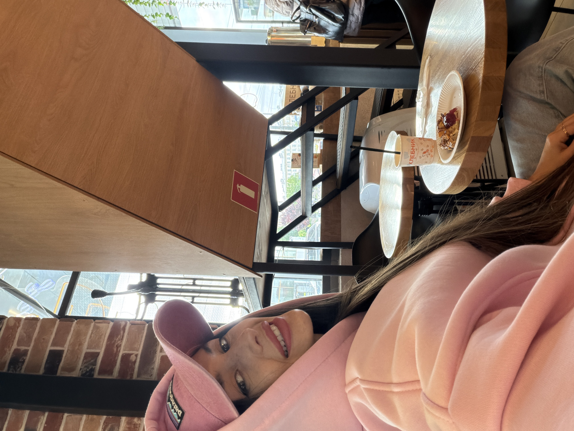 The image shows a woman in a pink hooded sweatshirt and a pink cap posing in what appears to be a cafe or coffee shop. She is smiling at the camera. In the background, there is a small round wooden table with a cup and a plate with a dessert on it. The cafe has an industrial-style interior with exposed brick walls and a staircase with black metal railings. Through the windows, a glimpse of the city is visible, potentially placing this location in Saint Petersburg, Russia, given the Cyrillic text on the cup which reads "ХЛЕБНИК". There's also a fire extinguisher sign on a wooden panel.
