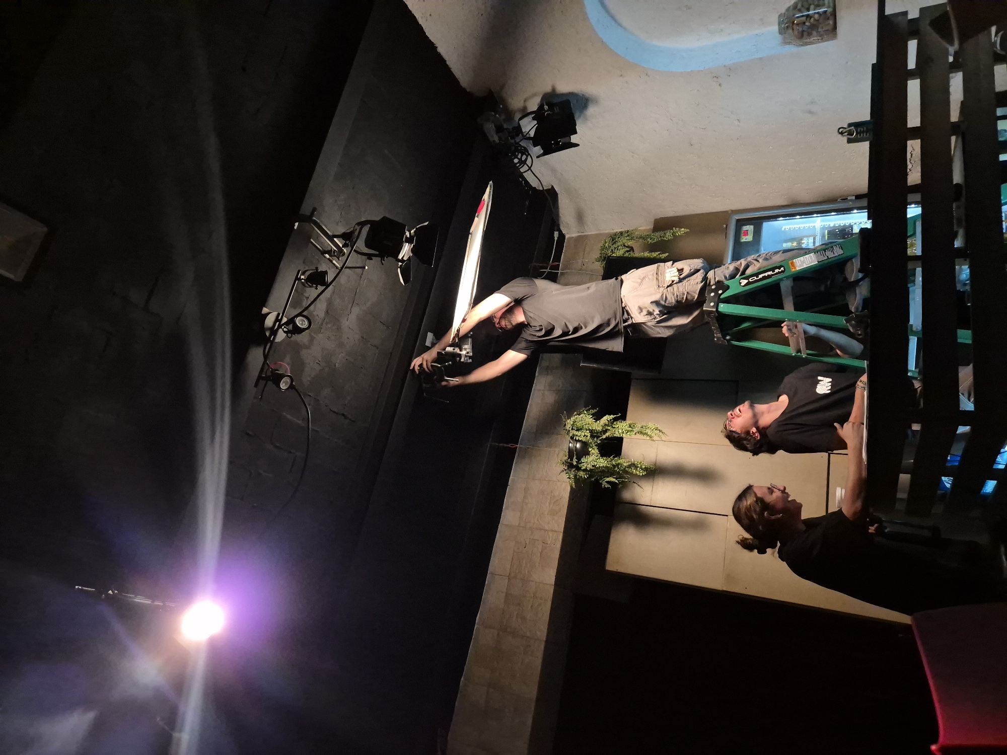 The image depicts a film shoot in what appears to be an interior space in the city of Puebla, Mexico. The set seems to be an industrial or commercial space with exposed brick or concrete walls. There are at least three people present. One man is standing, facing a camera and an arclight. Another person is standing on a Cuprum ladder, operating some kind of equipment. A third person is on the ground, also seemingly engaged in the filming process. The lighting setup consists of lights on stands. The set decoration includes greenery in planters. The background reveals elements of the location, including a refrigerated display case.