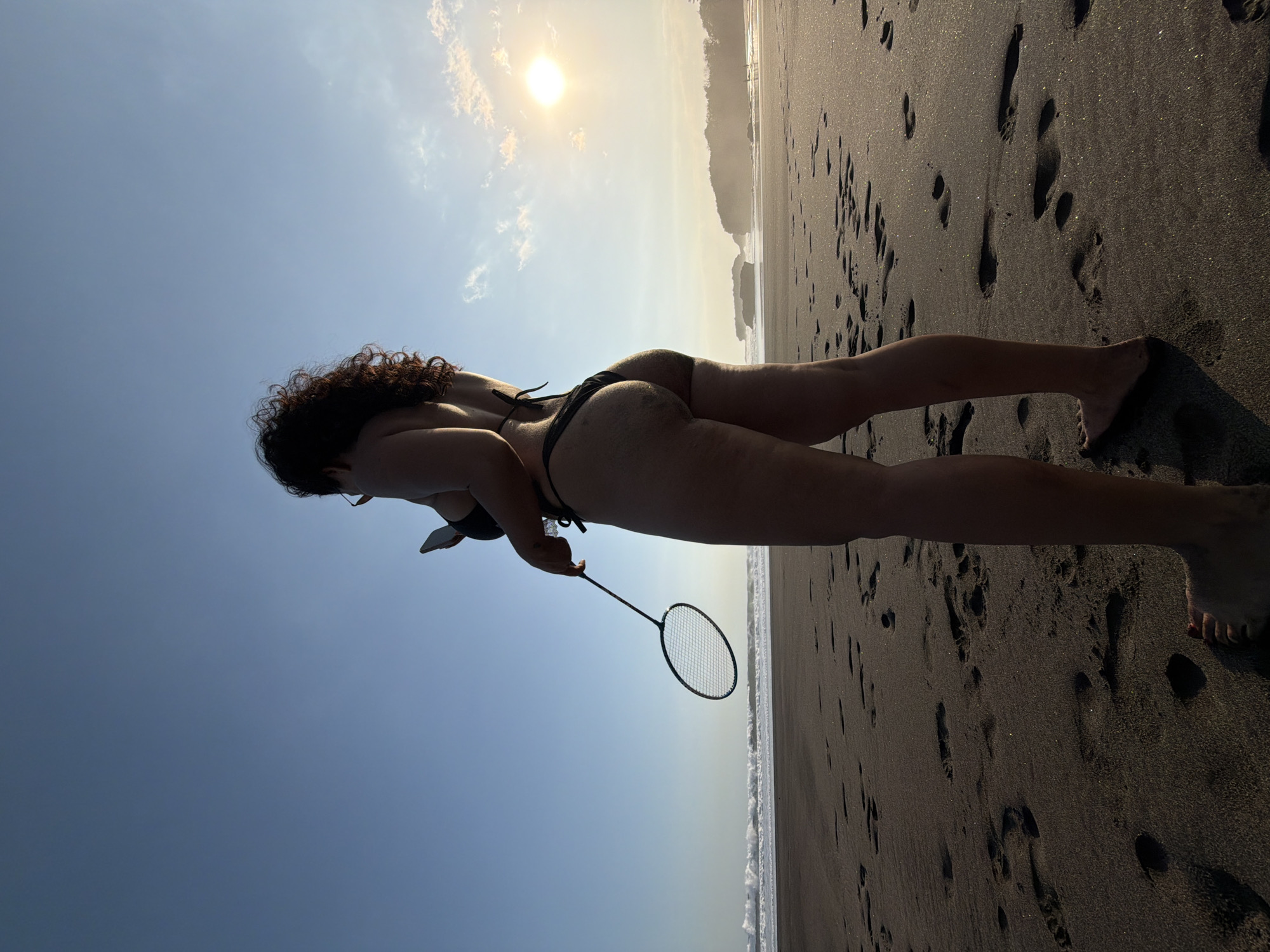 An outdoor, eye-level full shot captures a person standing on a dark sand beach during sunset in Selemadeg, Indonesia. The individual, depicted from a rear-right perspective in a semi-silhouette against the bright sky, appears to be a female with curly, dark hair. She is wearing a dark two-piece swimsuit, with sand visible on the back of the bikini bottom. Her left hand holds a badminton racket, while her right hand holds a smartphone, partially obscured. The beach features numerous footprints in the dark sand. To the left, ocean waves break with white foam. In the mid-ground, to the right, large dark rock formations or distant land are visible. The sun, a bright circular disc, is low in the upper right quadrant of the frame, indicating the late afternoon or early evening. The sky is largely clear blue, with a few faint clouds near the horizon.