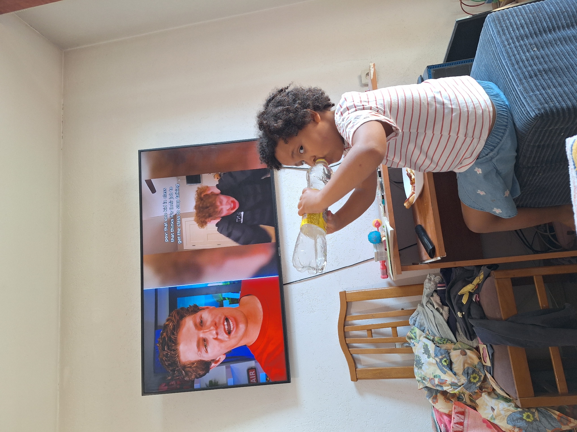 A young child with dark, curly hair is seated on a dark blue ottoman, drinking from a clear plastic bottle of water. The child is wearing a white t-shirt with thin red stripes and a light blue skirt with white floral patterns. In the background, a large flat-screen television displays a video with two men. The man on the left is wearing a red t-shirt and has brown hair. The man on the right, in the smaller portion of the screen, has red hair and is wearing a black hoodie. Text overlays are visible on the television screen. To the left of the television is a wooden chair piled high with various clothing items and blankets. To the right of the ottoman, a wooden desk is visible, with a remote control and a plate with food on it. The walls are painted a light color and appear to be textured. The scene is indoors, likely a living area. The location is not specified as Blagoevgrad, Bulgaria.