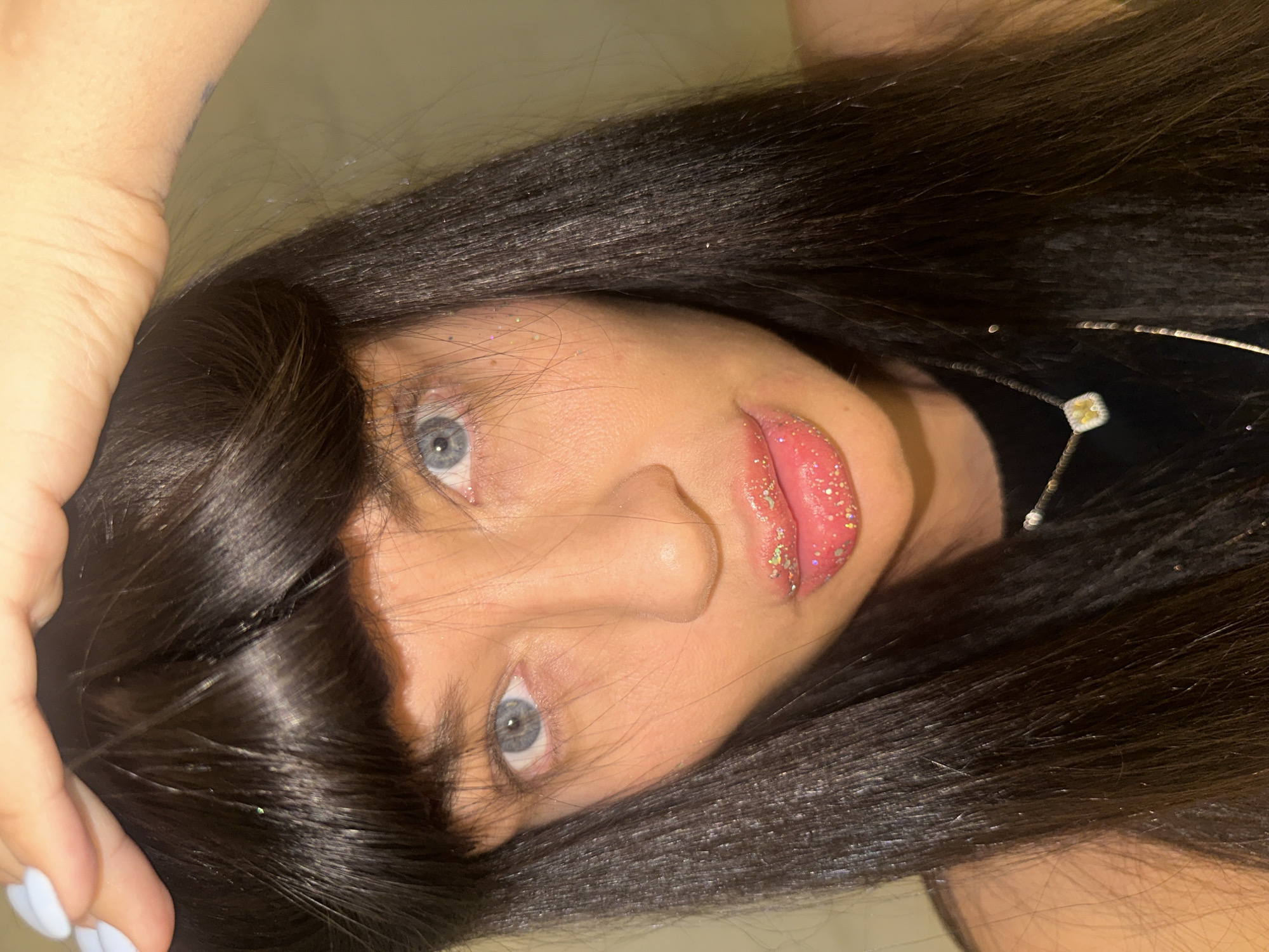 This close-up portrait features a person, likely a woman, with long, straight, dark hair cascading around her head. The image is taken from an overhead perspective, with her face tilted upwards, revealing her direct gaze with blue eyes. Her full lips are adorned with glossy, glittery pink lipstick, and iridescent sparkles are noticeable on her lips and scattered throughout her dark hair.
A thin silver necklace with a small, light-colored, possibly yellow or white, pendant is visible against a dark top worn by the subject. On the left side of the frame, part of a hand and forearm are visible, with the fingertips showing light blue painted nails. A faint marking or tattoo can be seen on the inner arm. The background is an indistinct, plain, light-colored surface, indicating an indoor setting. The subject maintains a neutral expression, engaging directly with the camera. The overall impression is a stylish portrait, highlighting makeup and hair details. No text is visible in the image, which is set in Moscow, Russia.