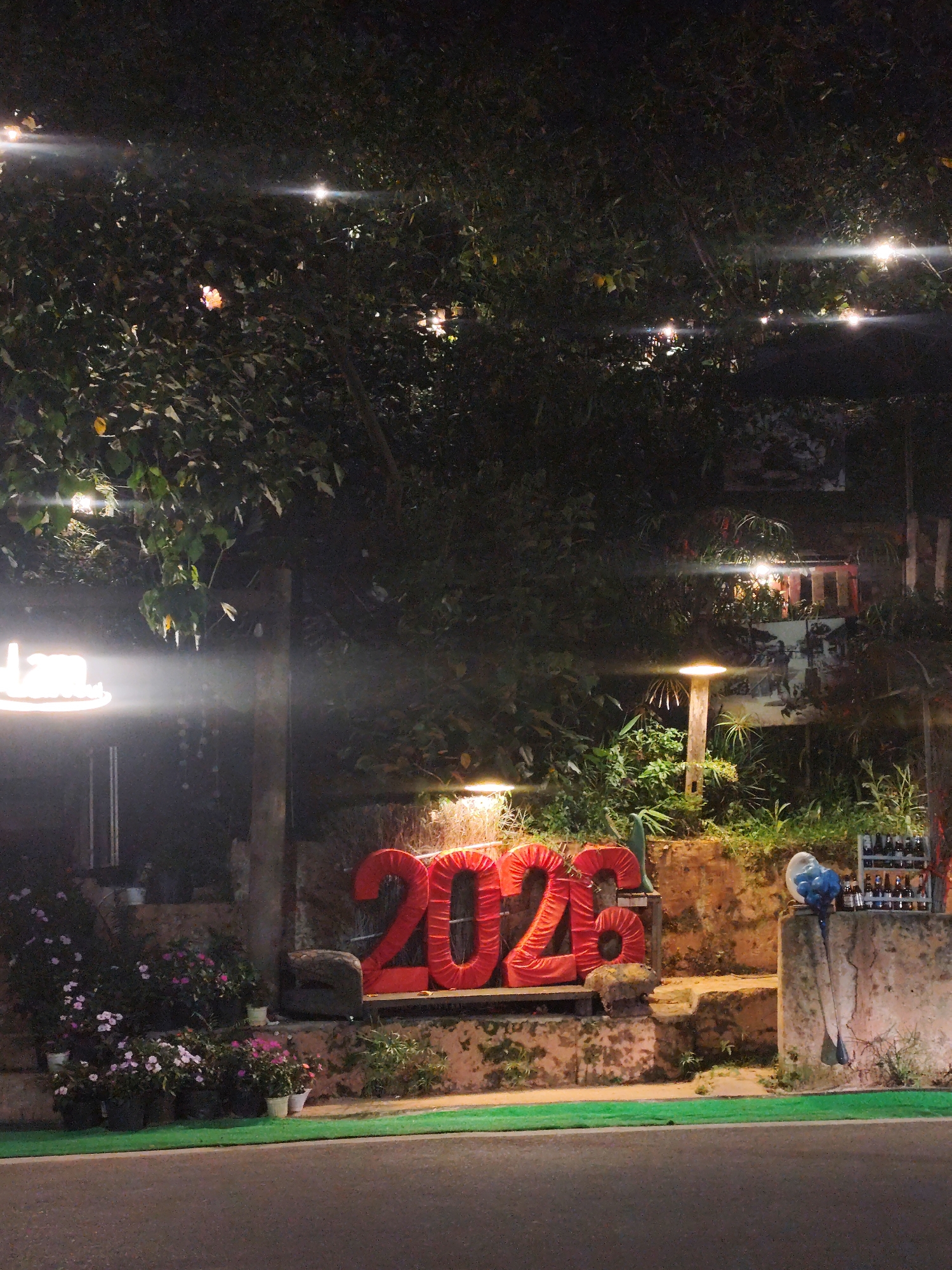 At night, in Da Lat, Vietnam, a roadside display features red, inflated numbers "2023" on a stone ledge. A selection of drinks is displayed to the right of the numbers, and various plants and decorative elements frame the scene. Overhead, strings of lights illuminate the trees above the display.