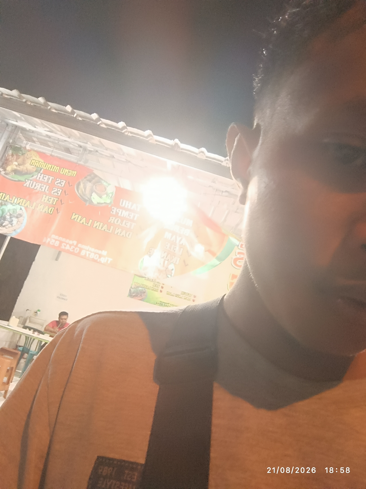 The image, taken on August 21, 2026, at 18:58, captures a selfie of an individual in the foreground, with a blurred background that suggests an outdoor setting possibly in Sidodadi, Indonesia. A banner with text in Indonesian is visible, indicating menu items such as "ES TEH" (iced tea) and "AYAM TETE" (chicken tetes). The banner also displays a phone number: "0812 8374 9924". In the middle ground, a person in a red shirt is seated at a table. The overall environment appears to be a street-side food stall or a casual dining area, illuminated by artificial light.