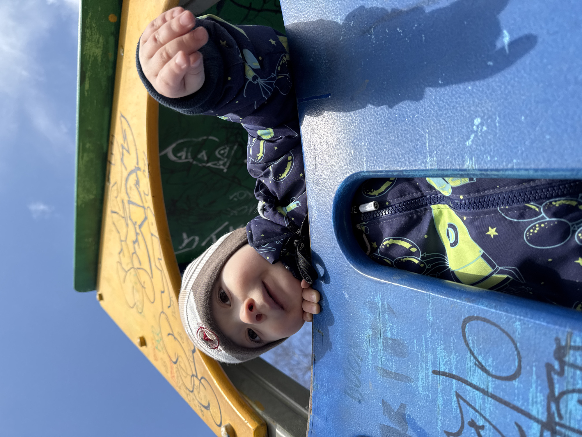 A young child peeks down from a brightly colored playground structure, captured from a low-angle perspective in Izhevsk, Russia. The child, likely a toddler, wears a dark blue winter jacket patterned with light green rockets and yellow stars, and a beige and white striped beanie. Their left hand is visible, resting on the yellow panel of the structure. The child's face, with wide, curious eyes and a slightly open mouth, looks directly towards the viewer, conveying a sense of playful engagement.
The playground structure consists of a yellow panel on the left with faint, dark, abstract graffiti, and a textured blue panel on the right, partially obscuring the child's body. This blue panel also features more distinct black and blue graffiti, including stylized letters and tags. Bright sunlight illuminates the scene, casting strong shadows on the blue surface, indicating a clear, sunny day. A vibrant, cloudless blue sky fills the upper left background. The image captures a moment of childhood exploration in an outdoor setting.