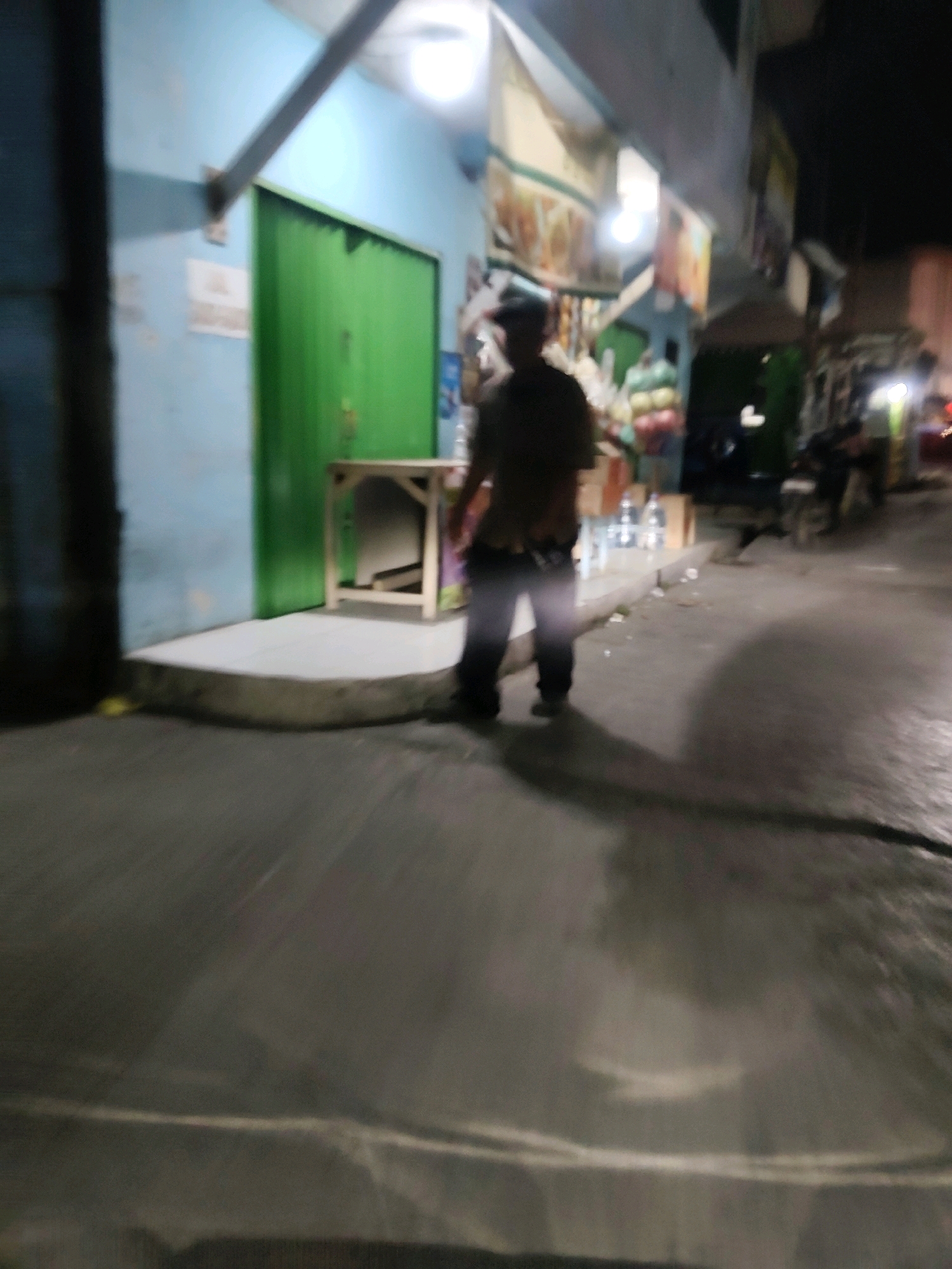 The image shows a blurry, nighttime scene in front of a building with a light blue facade and a green door. A person, silhouetted and out of focus, stands on a sidewalk in front of the building, wearing dark clothing. To the right of the person, a small wooden table is visible. In the background, there are some goods displayed on shelves and possibly a motorcycle parked. The street in front of the building appears wet, reflecting the lights. The overall impression is that of a small shop or roadside stall in a dimly lit urban environment. This location is identified as Mekarmukti, Indonesia.