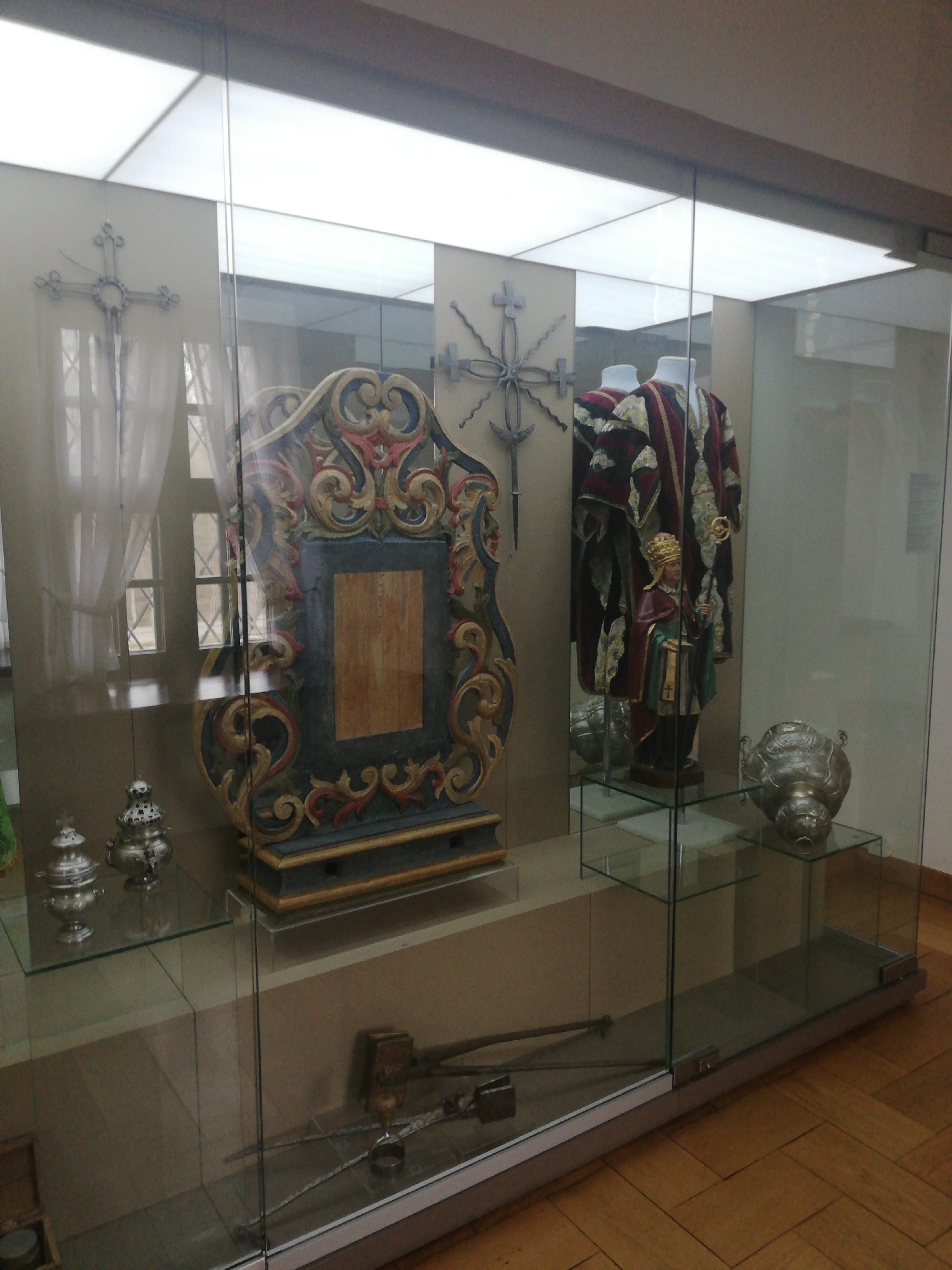 This image depicts a museum exhibition hall in the city of Nyasvizh, Belarus, showcasing historical and religious artifacts within a large, multi-section glass display case.
The foreground is dominated by a clear glass display case with a light brown base. The floor is constructed of light-colored wooden planks arranged in a parquet pattern. The ceiling features several large, illuminated rectangular white panels.
Inside the leftmost section of the display, a decorative metal cross, possibly wrought iron, is mounted on a light-colored wall. Below it, on a clear glass shelf, two ornate silver-colored metal vessels with lids are exhibited. Behind these objects, a window with a grid pattern and sheer white curtains is partially visible, either directly or as a reflection on the glass.
The central section features a large, elaborately carved wooden frame, painted predominantly in blue, gold, and red/pink hues, standing upright. Its central panel is a recessed, plain wooden surface.
In the rightmost section of the display, a mannequin is dressed in a rich ecclesiastical vestment, characterized by patterns in red, gold, and dark fabric. In front of this mannequin, a smaller, carved wooden statue of a figure, possibly a cleric or saint, wearing a gold crown or mitre and holding a staff, stands on a dark pedestal. To the right of the statue, on a separate clear glass shelf, a large, ornate metal vessel, resembling a censer or urn, rests on its side.
On the lowest level of the central display, inside the glass case, several antique metal tools or implements, including what appears to be a hammer and tongs, are laid out on a beige surface.