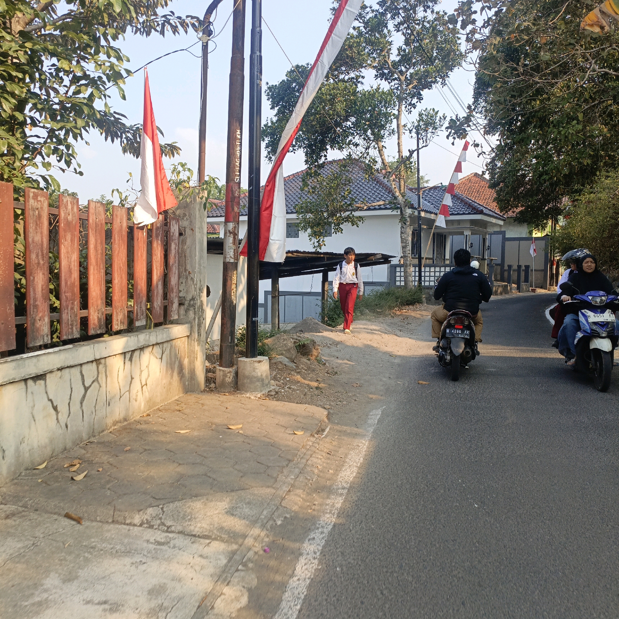 The image depicts a street scene in Salatiga, Indonesia, with a clear sky. In the foreground, a paved road with a white line marking the edge is visible. Two motorcycles are traveling on the road. The motorcycle on the right carries two people, a woman driving and a passenger behind her. The motorcycle on the left has a single rider. To the left of the road is a sidewalk and a wooden fence. A young person in a white shirt and red pants, likely a student with a backpack, is walking on the sidewalk. In the background, a white building with a tiled roof is situated behind the fence. Several Indonesian flags, red and white, are displayed on poles and along the fence. The surrounding area features trees with green foliage and a concrete retaining wall with a textured surface. The lighting suggests it is either early morning or late afternoon.