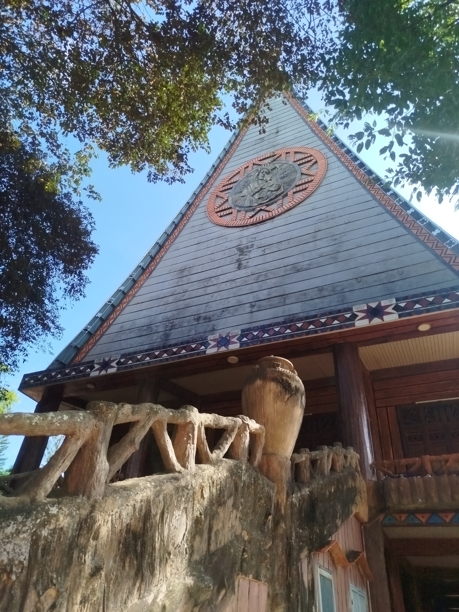 This is a ground-level shot of a building in An Phu Ward, Vietnam. The building features a triangular facade made of gray wood planks, decorated with geometric patterns along the edges. A circular emblem is set into the triangle near its apex. Below the facade is a wooden eave decorated with star-shaped patterns. The facade is supported by dark brown wooden columns.
In the foreground, there is a textured cement railing with a large vase-shaped cement post. The railing appears to lead up to the building's entrance. Some tree branches with leaves are visible in the top portion of the frame, framing the building and sky. The sky is a clear blue. The shot is taken in daylight.