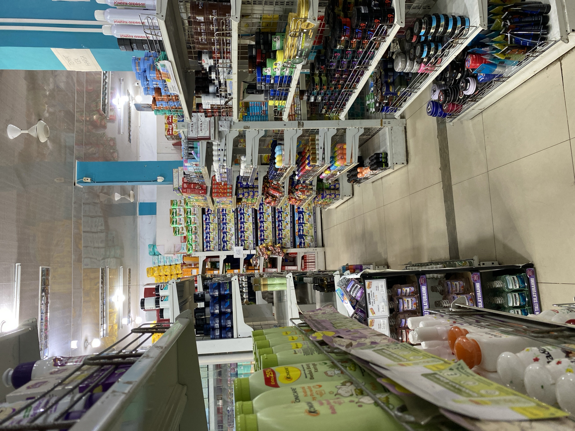 The image shows the interior of a well-lit supermarket aisle. The shelves are stocked with various products, primarily personal care items and household goods. On the left, shelves display green bottles of what appear to be baby powder or lotion, with labels like "OniCARE." Further back, there are rows of yellow and white packaged goods, possibly diapers or sanitary products, and various canned and boxed items.
In the center of the image, shelves are filled with numerous small containers of hair styling products, such as gels and pomades, in blue, black, and silver packaging. There are also bottles of mouthwash, specifically Listerine, displayed in the lower right section. To the right, larger bottles of personal care products, possibly shampoo or conditioner, are visible. The flooring is tiled and a ceiling fan is visible overhead. Signs are present, including one with text that appears to be in Indonesian, possibly indicating prices or product information. The overall impression is that of a typical retail environment in a small to medium-sized town. The location is identified as Lampisang, Indonesia.