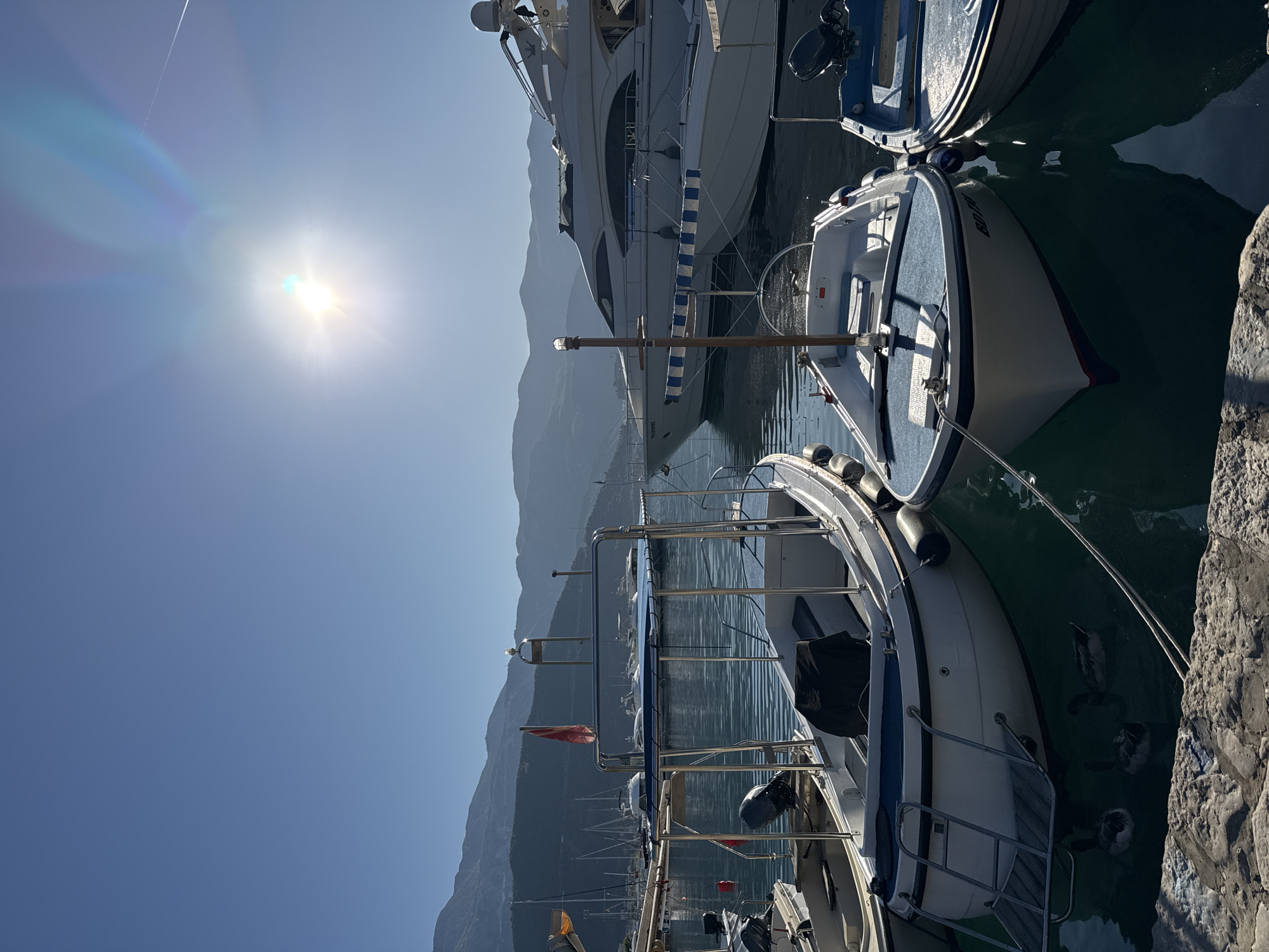 The image depicts a marina in Budva, Montenegro. Several boats of varying sizes are docked close together in the water. A large yacht is partially visible, along with smaller fishing boats. A mountain range is visible in the background, silhouetted against a bright, sunny sky. The sun is positioned high in the sky, creating a glare. Stone structures appear on the right edge of the frame, likely forming part of the harbor's infrastructure. The water is calm, and details of boat rigging and other fixtures are visible.