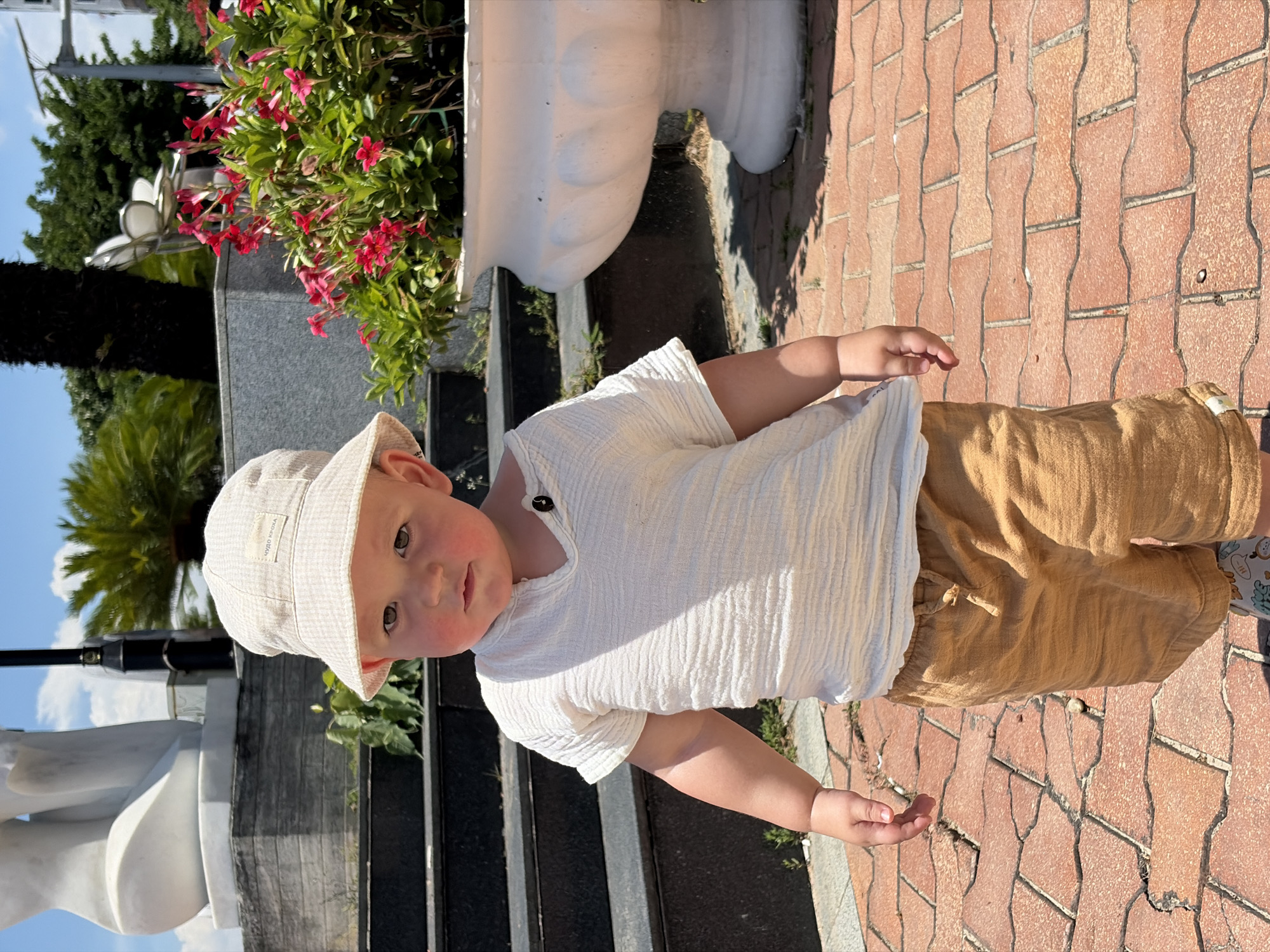 A small child with a beige bucket hat, wearing a white, short-sleeved shirt and brown shorts, stands on a brick pavement. The child is near a large white flower pot filled with red flowers and green foliage. Steps lead up behind the flower pot to a dark, possibly stone or granite, surface. In the background are trees and a partial view of a white sculpture. The pavement is made of red, brick-like interlocking pavers. Based on the visible architecture and landscaping, the location is likely a park or garden setting in Đà Nẵng, Vietnam.