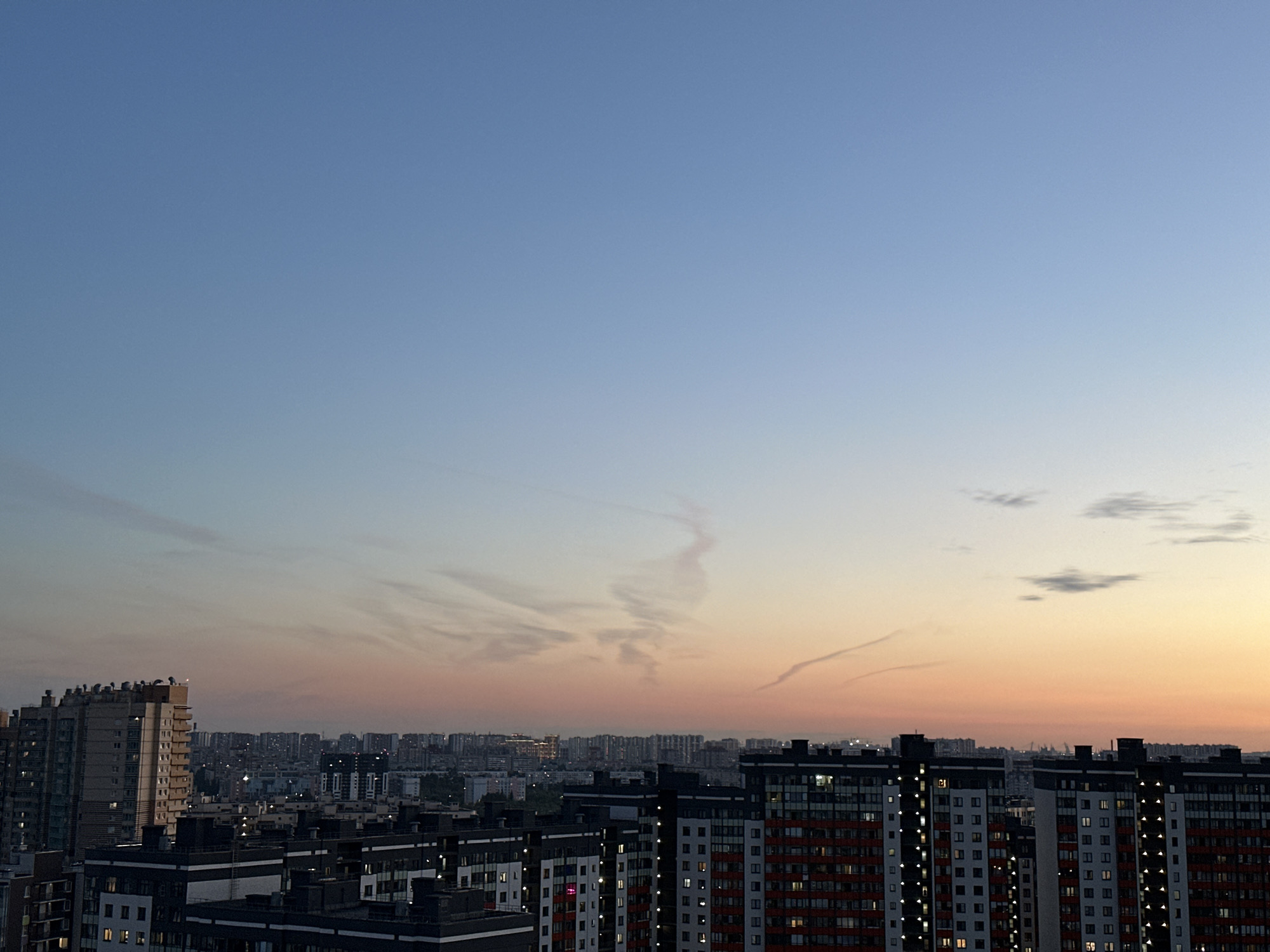 The image captures a wide-angle, high-angle view of a cityscape at dusk in Otradnoye, Russia. The sky transitions from a clear, pale blue at the top to warm hues of pink and orange near the horizon, indicating the setting sun. Wispy, light-colored clouds are scattered across the sky, with a more prominent, elongated cloud formation visible in the mid-sky.
In the foreground, a series of modern, multi-story apartment buildings are prominent. The building on the far left is a tall, light-colored structure with many windows. To its right and stretching across the frame are several darker buildings with visible windows illuminated from within, suggesting occupancy. These buildings feature a mix of white, gray, and red facade elements.
In the background, a dense urban landscape stretches to the horizon, with numerous buildings of varying heights. The distant city lights are visible, creating a subtle glow against the twilight sky. The overall environment appears to be a densely populated residential area. There are no discernible people or specific actions depicted in the image. The scene is static, focusing on the architectural landscape and the atmospheric conditions of the sky.