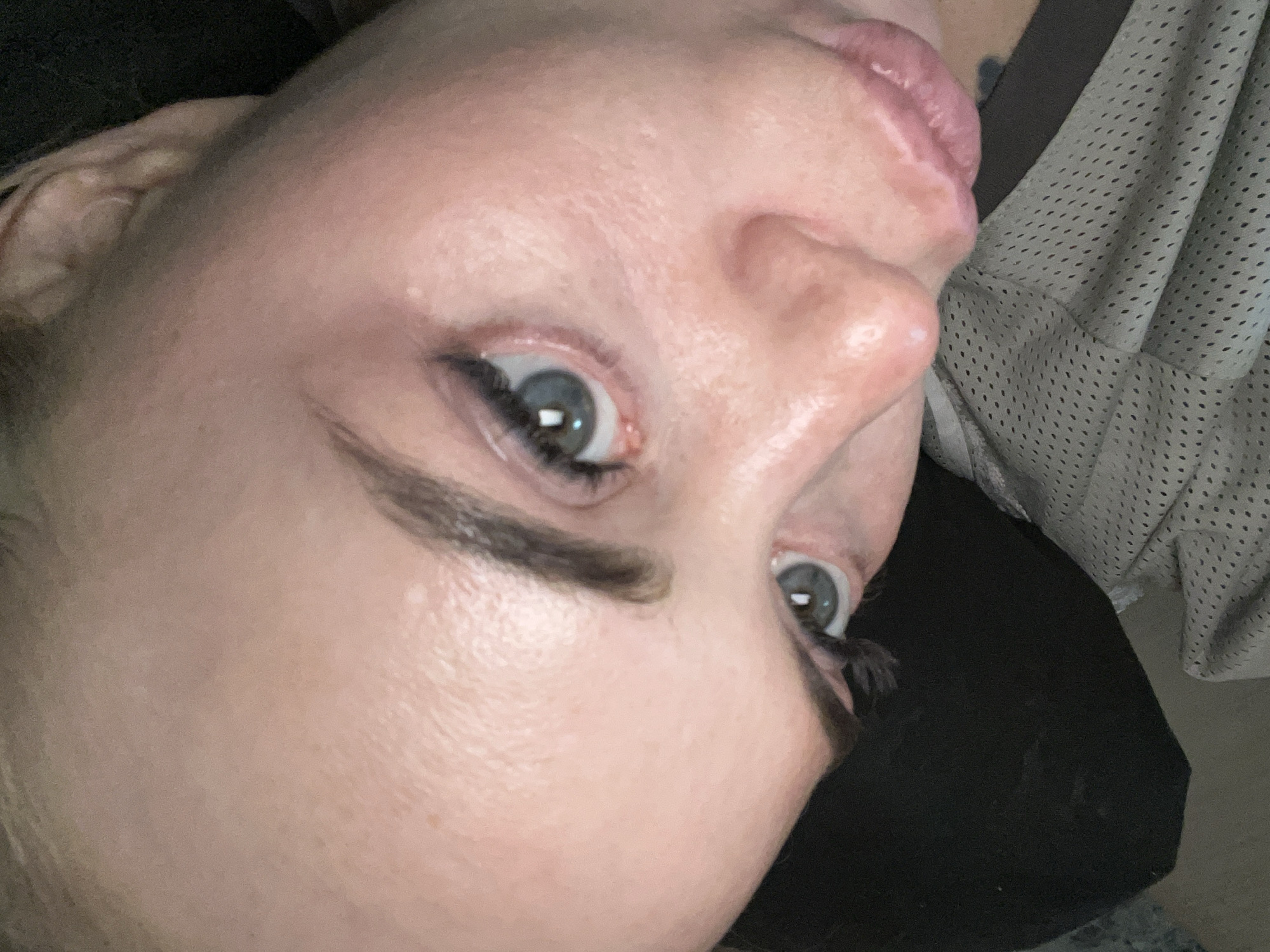 A close-up, overhead shot shows a person's face, tilted to the right. The person has light-colored skin, light blue eyes, and dark eyebrows. The left eye is in focus, with visible eyelashes and eyeliner. The person's lips are slightly parted. A portion of the person's forehead and nose are also visible. The background is dark, with a hint of textured fabric, possibly clothing. The image appears to be taken indoors.
