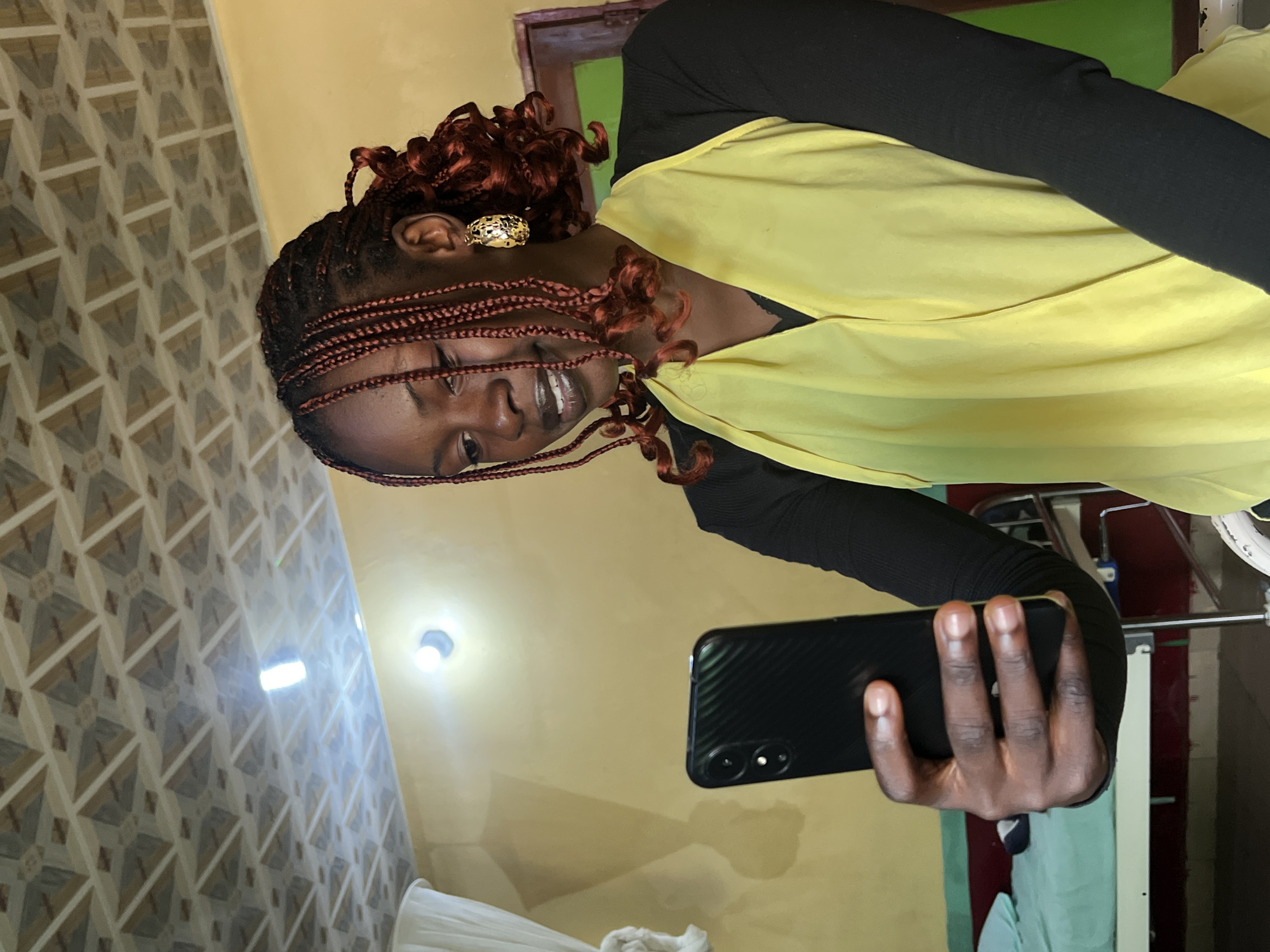 A young Black woman with braided hair and large gold hoop earrings is taking a selfie with her smartphone in an indoor setting. She is wearing a black long-sleeved shirt underneath a yellow wrap-style top. The background appears to be a room with yellow walls and a patterned ceiling. To the left, a hospital bed with a light green sheet is visible, suggesting the location could be a hospital or clinic in Jos, Nigeria. The lighting in the room is dim, with a bright light fixture on the ceiling.