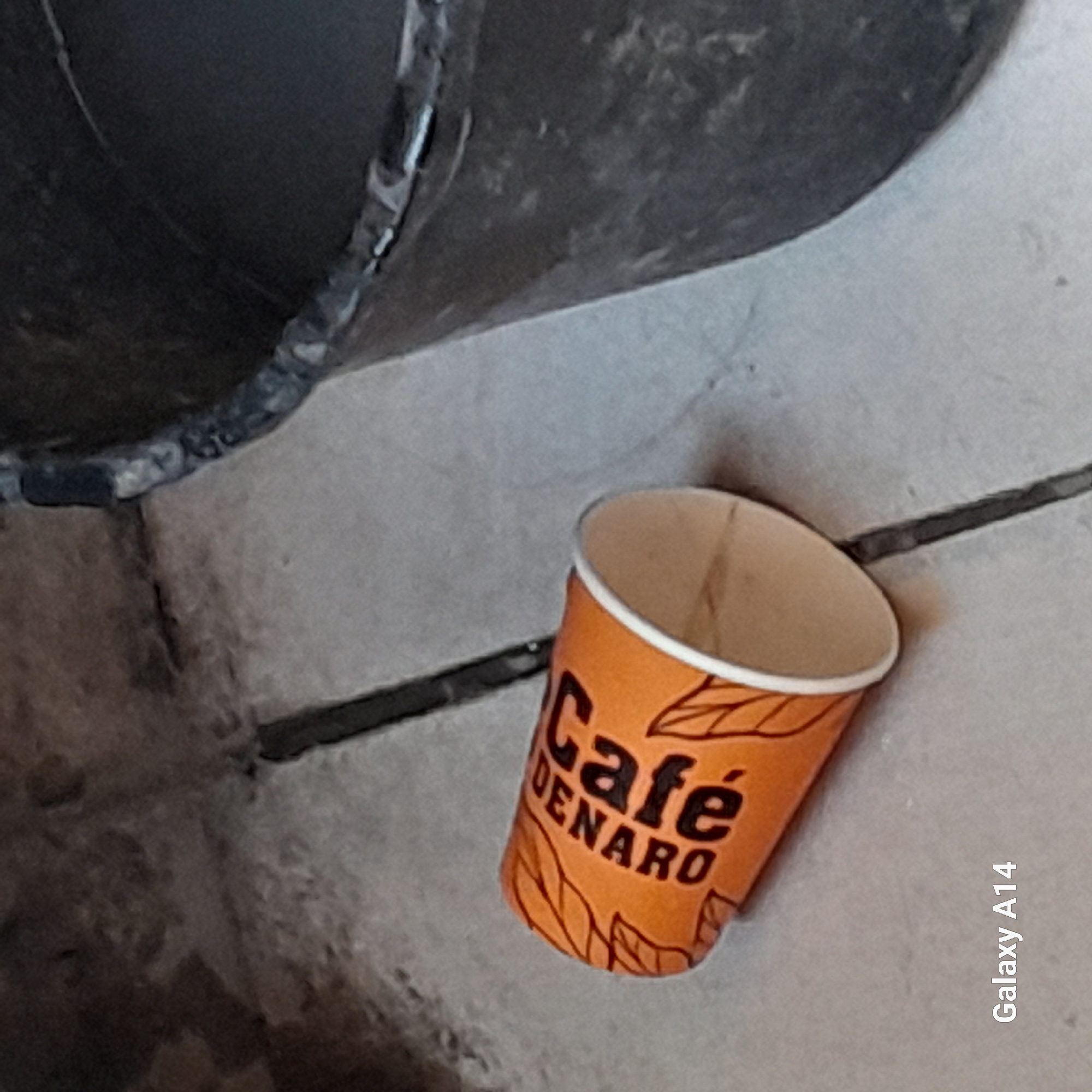 An orange disposable coffee cup with black lettering that reads "Café de Naro" and leaf motifs lies on a grey, tiled surface. A dark, textured cylindrical object, possibly a bucket, is positioned to the right of the cup. The background appears to be a rough, concrete-like texture. The bottom left corner of the image displays the text "Galaxy A14". The scene is indoors or outdoors in an urban setting, potentially in Khan Yunis, Palestinian Territories. There are no people visible in the image.